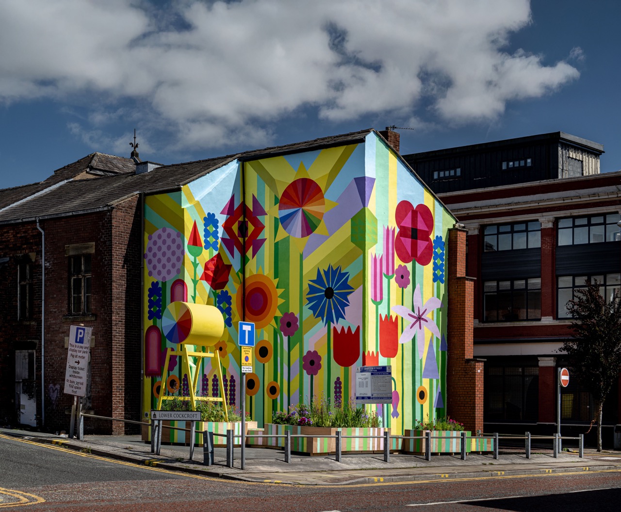 A vibrant mural with large colourful flowers, geometric shapes, and bold patterns covers the entire side of a building at a street corner under a partly cloudy sky. An art installation stands in front.