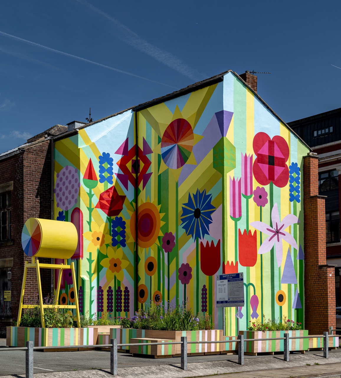 A vibrant mural covers the side of a building, featuring bold, geometric flowers in bright colours. A large yellow cylinder structure stands beside flower beds in front of the mural, all under a clear blue sky.