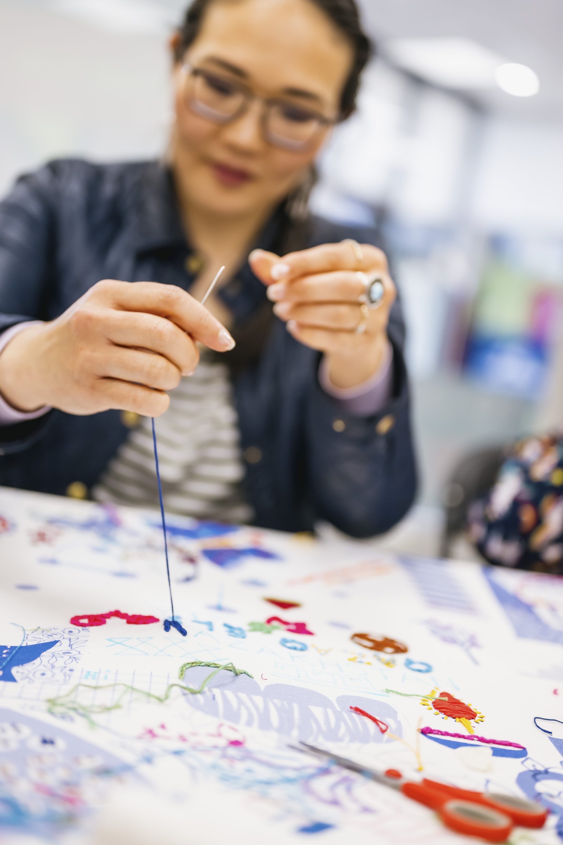 A woman with glasses is threading a needle over a colourful piece of fabric covered in various painted designs, working at a table in a bright, blurred indoor space.
