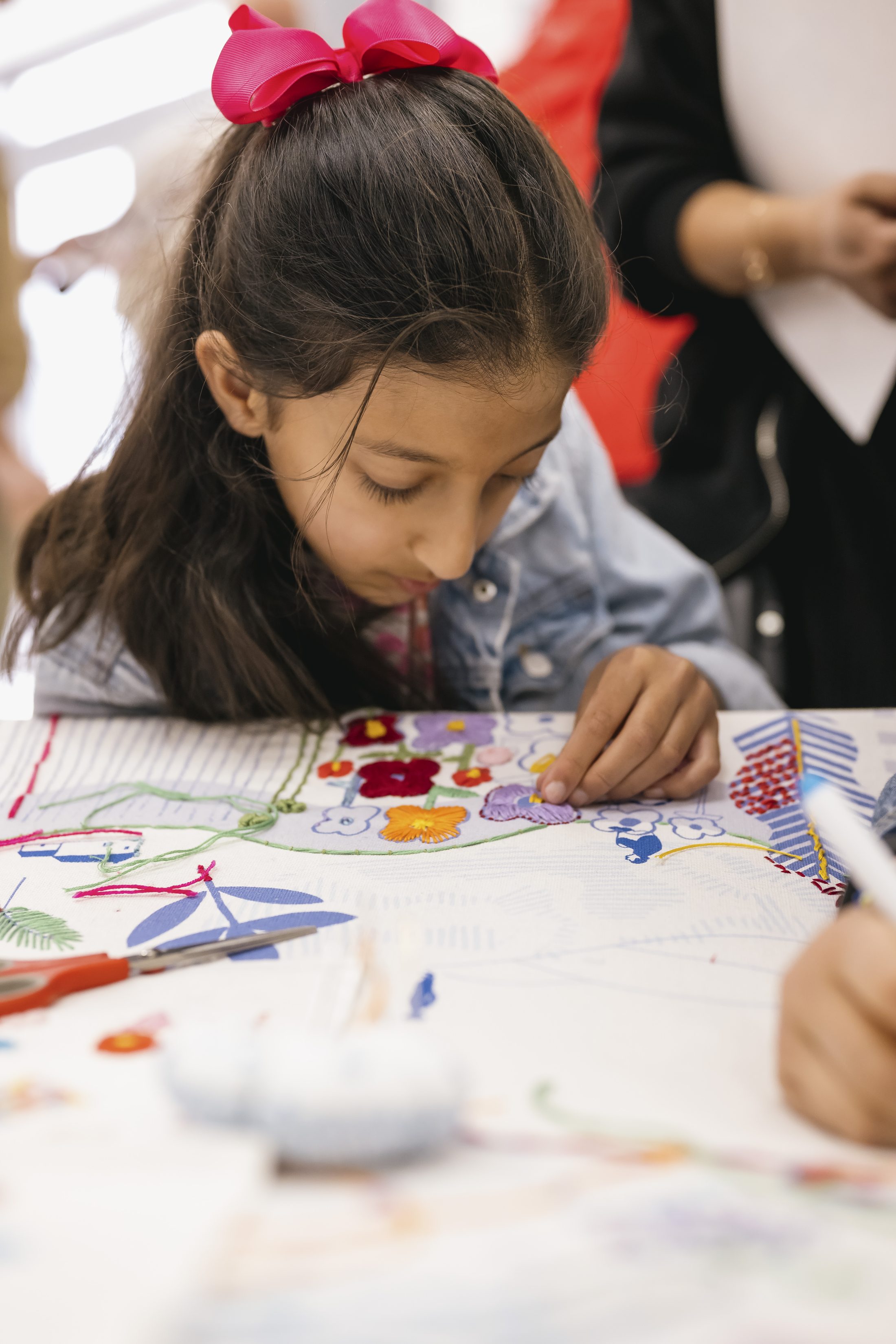 A young girl with a pink bow in her hair carefully works on a colourful embroidery project at a table, focussing intently on her stitching. Other people and art materials are visible in the background.