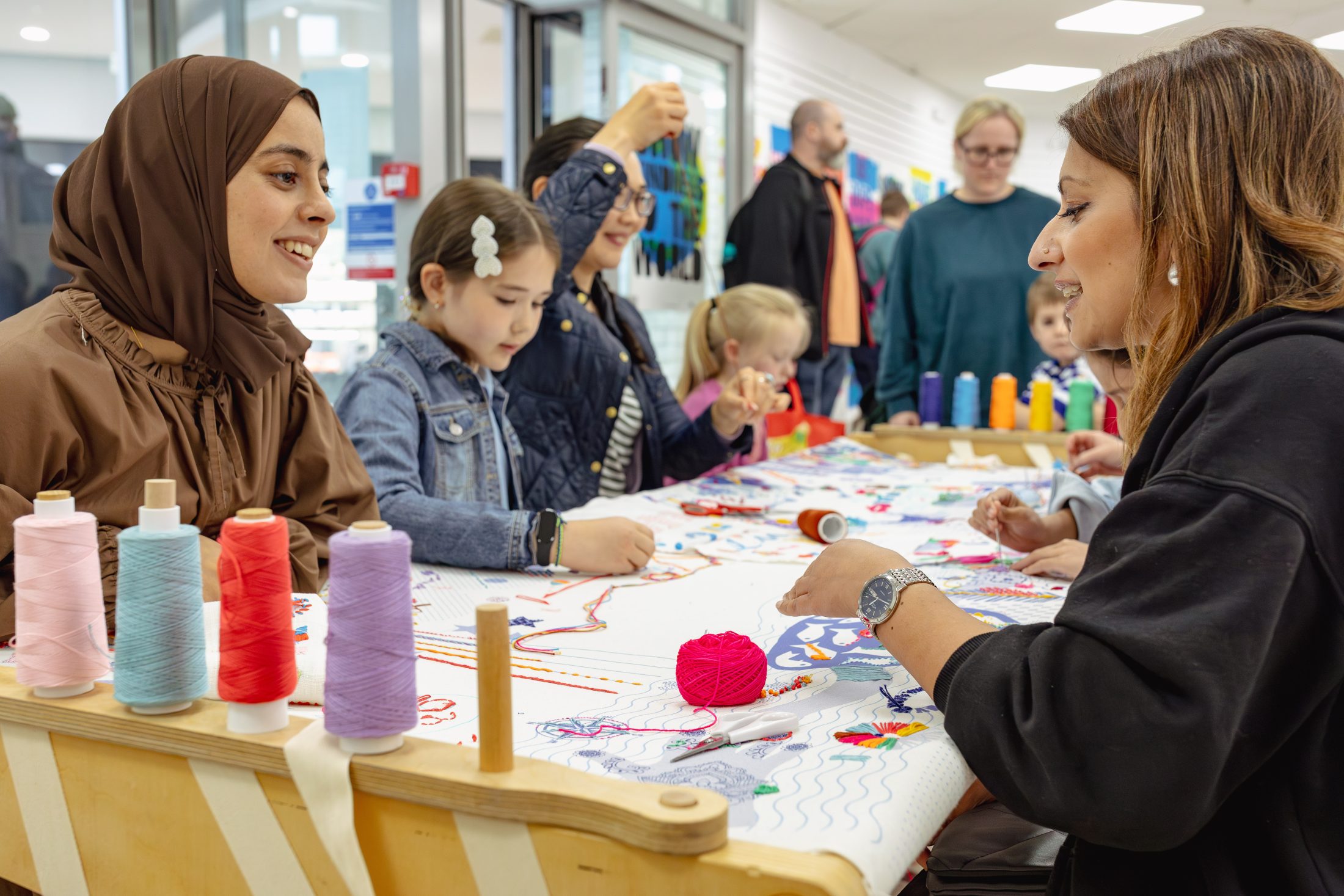 A group of women and children sit around a table covered in colourful threads, smiling and working on crafts together in a bright indoor setting. Various spools of thread are visible on the table.