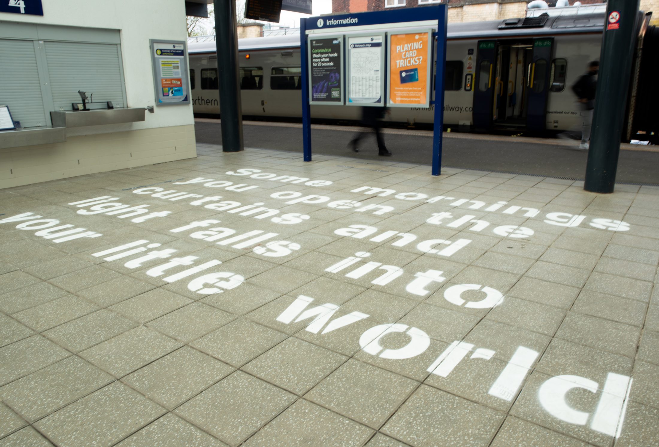 Text on the floor of a railway station reads: “Some mornings you open the curtains and the light falls into your little world.” A train and blurred figure are in the background near signs and ticket windows.