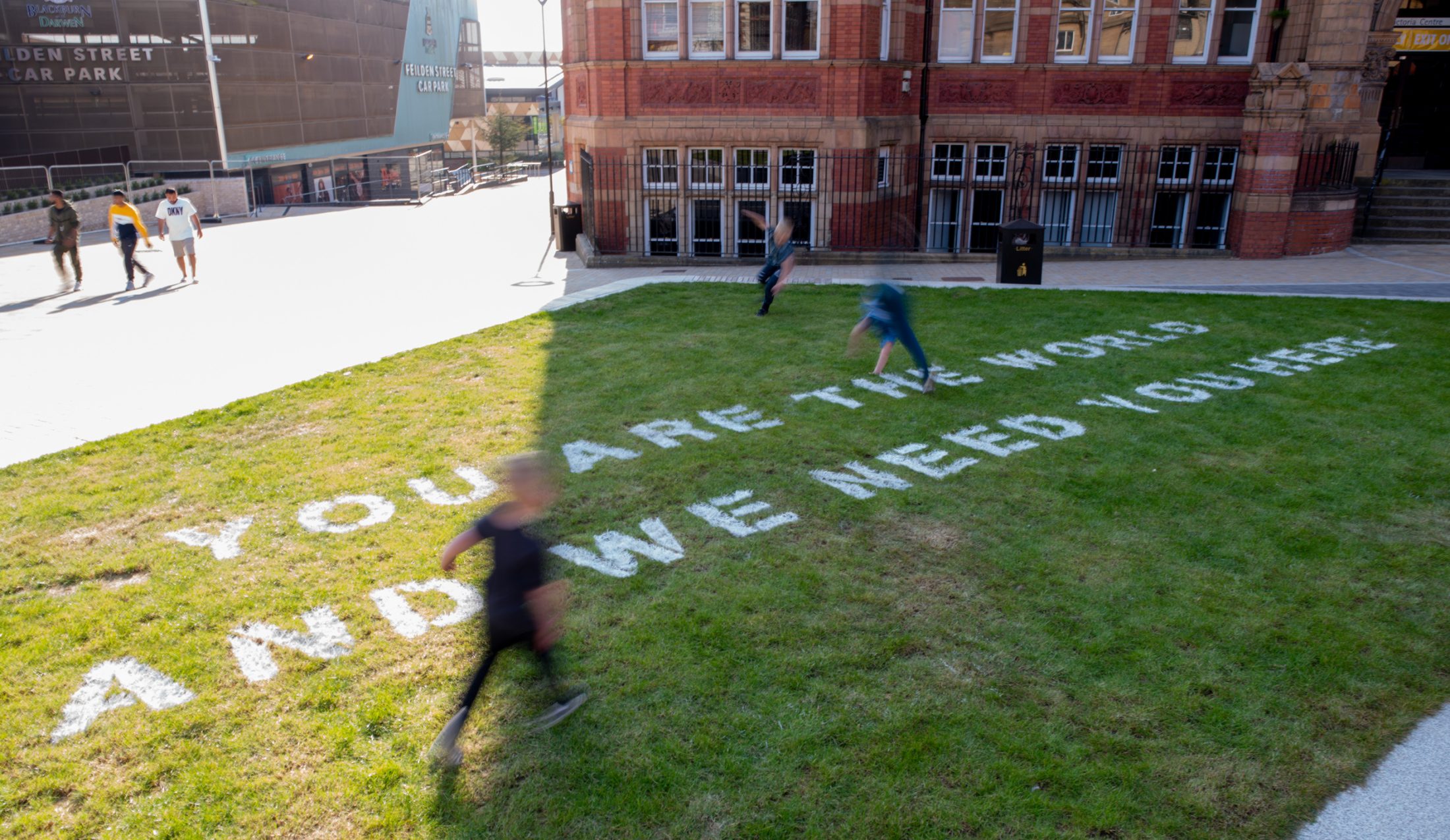 Grass lawn with white text reading “YOU ARE THE WORDS AND WE NEED YOU HERE” near a red-brick building, with blurred figures suggesting playful movement.