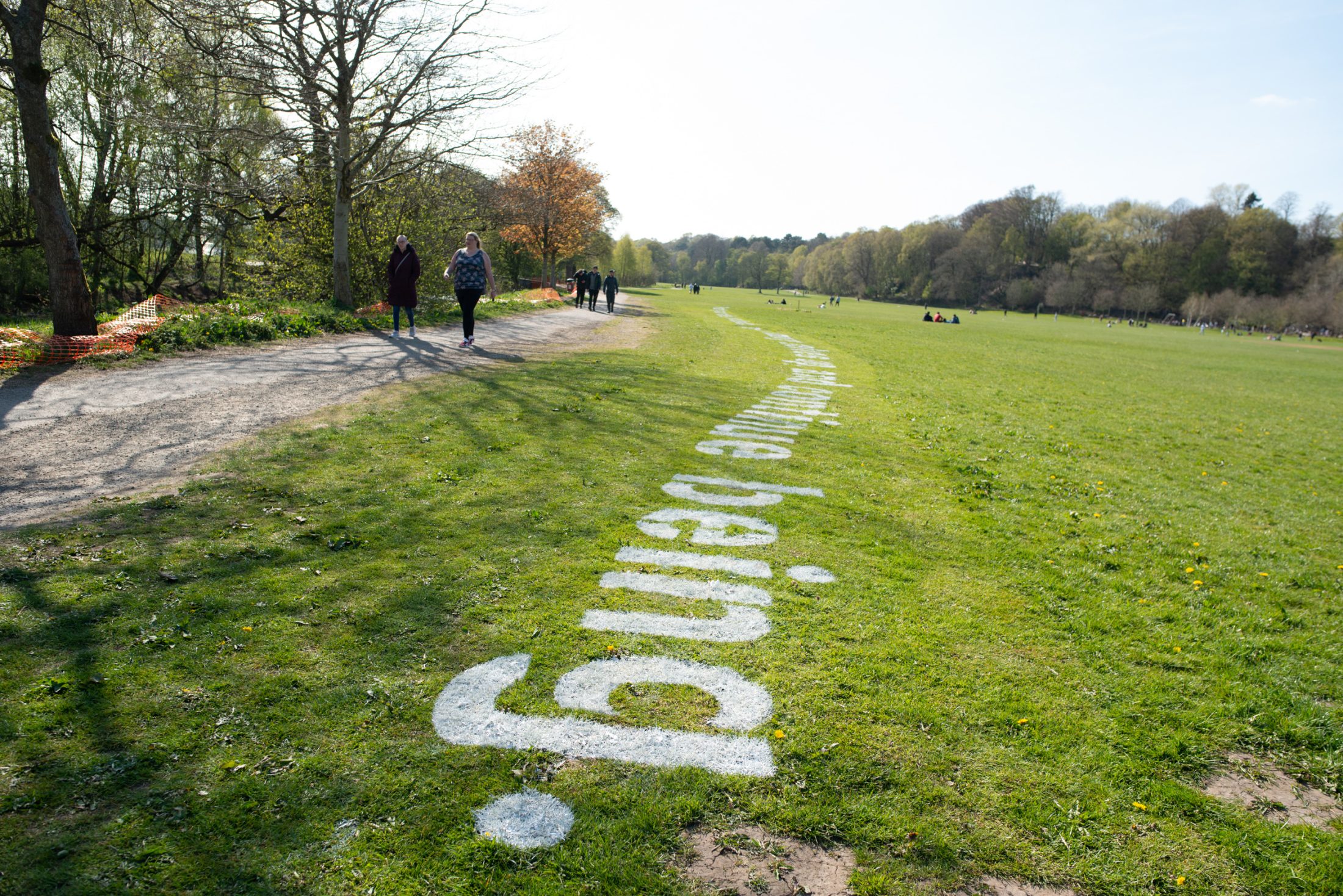 A park with people walking along a path; large white text reading time being. is painted on the grass beside the path, and trees and open green spaces are visible in the background.