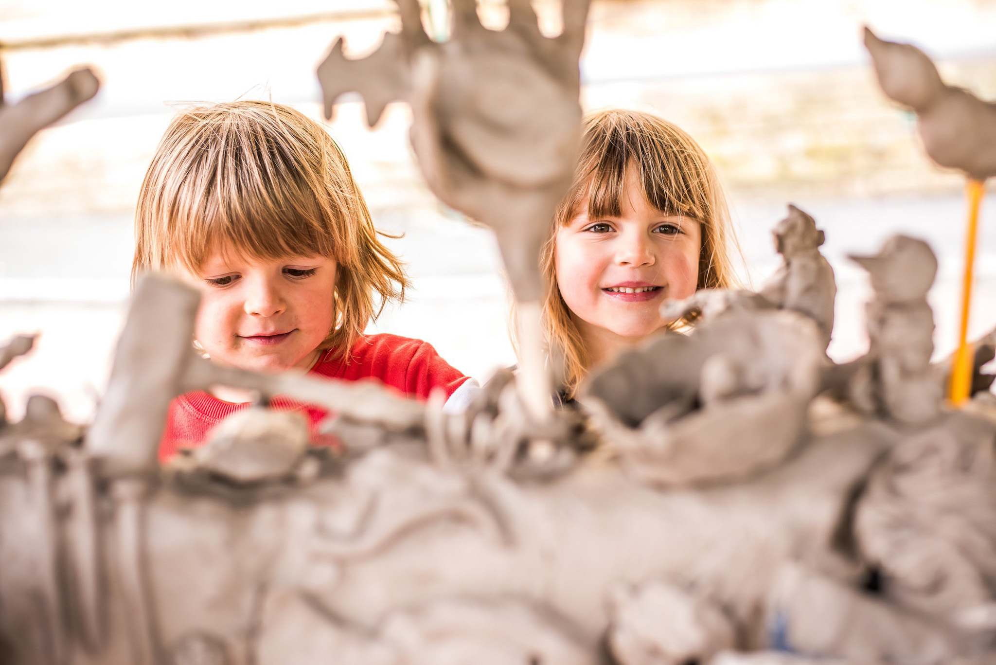 Two young children with light brown hair, one in a red shirt and one in a white shirt, smiling and playing with a large, messy sculpture made of clay or similar material. The scene is bright and cheerful.
