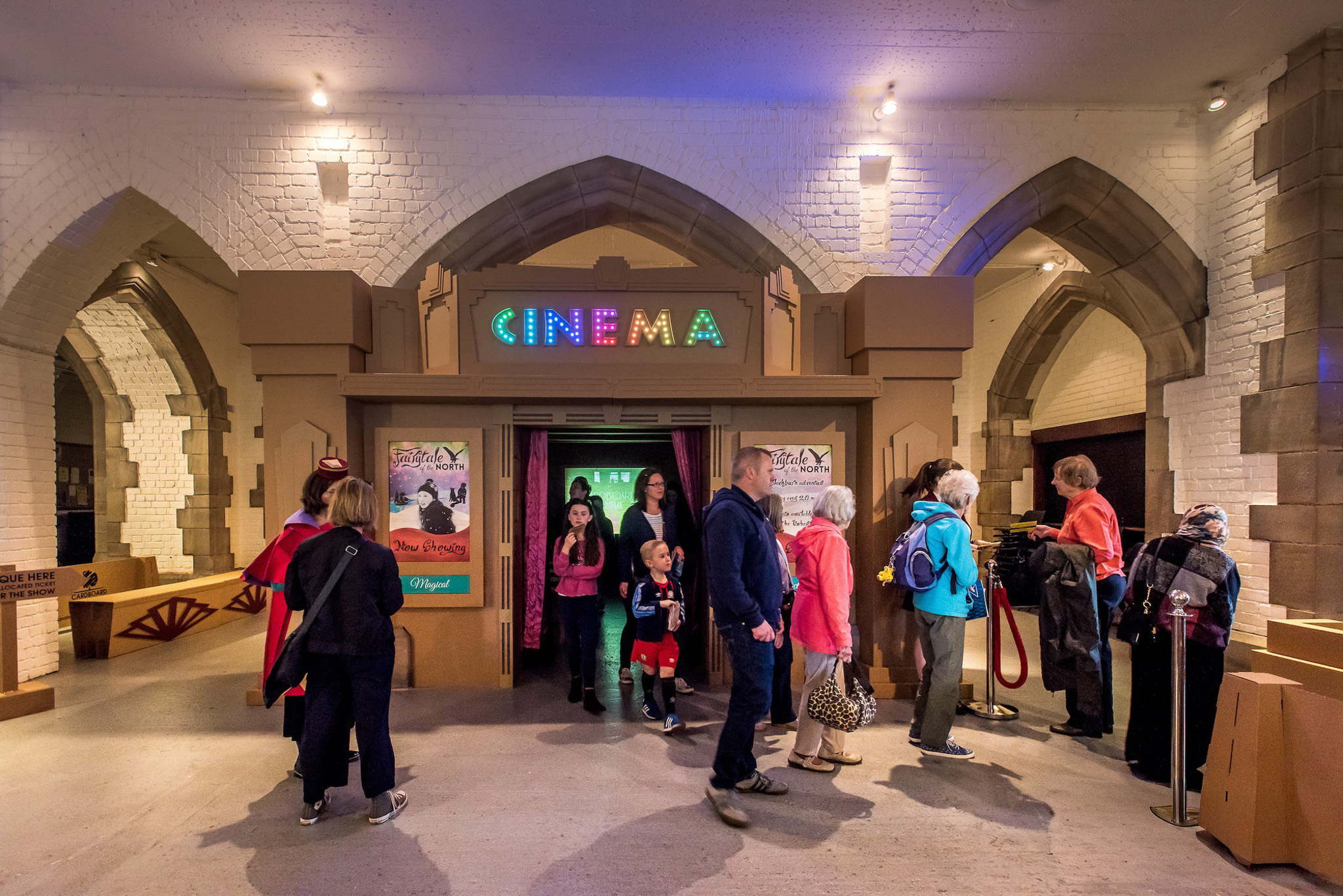 A group of people stands in front of a vintage-style cardboard cinema entrance in a stone archway. Lit letters spell 
