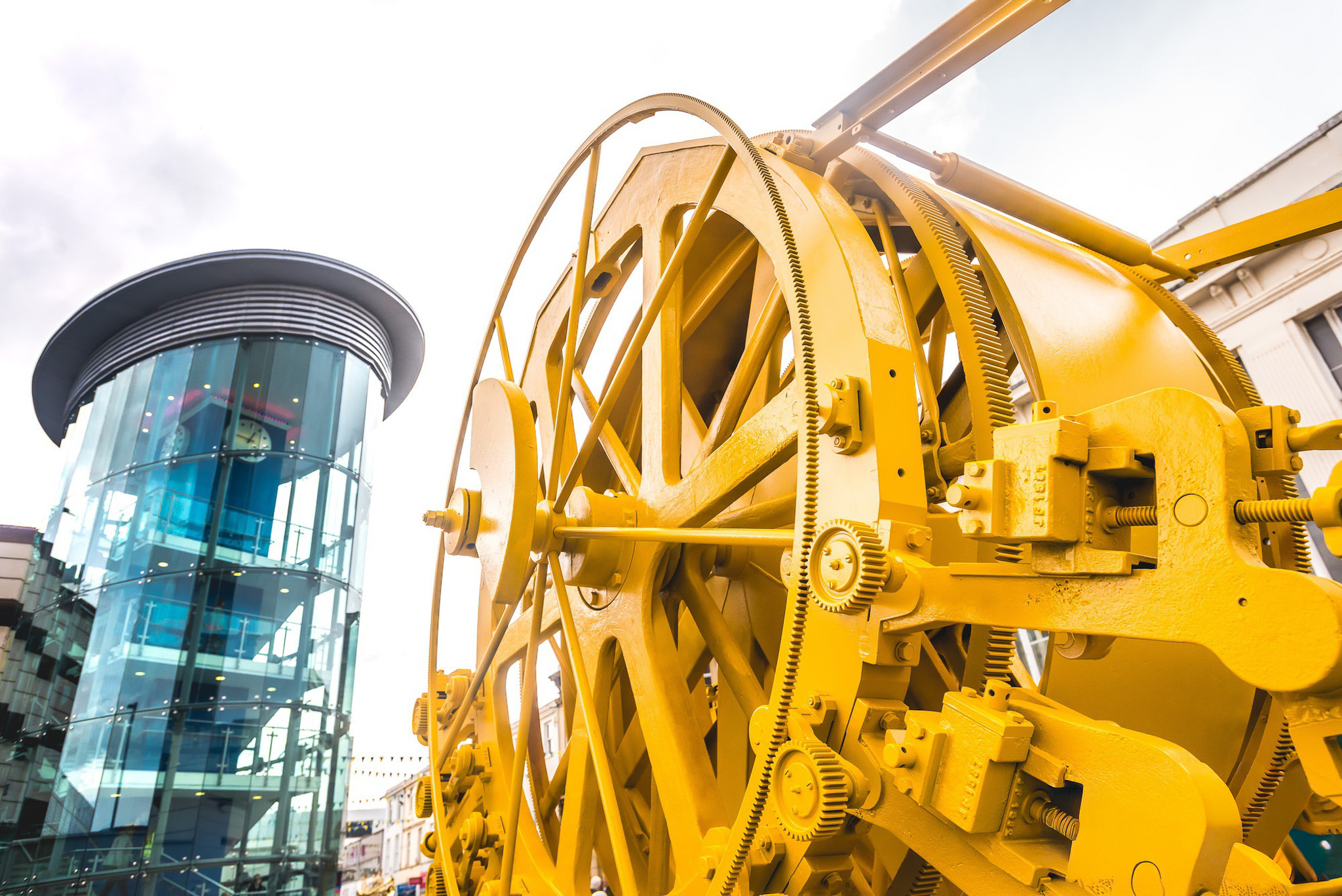 A large yellow industrial machine with gears is displayed outdoors near a modern glass cylindrical building under a cloudy sky.