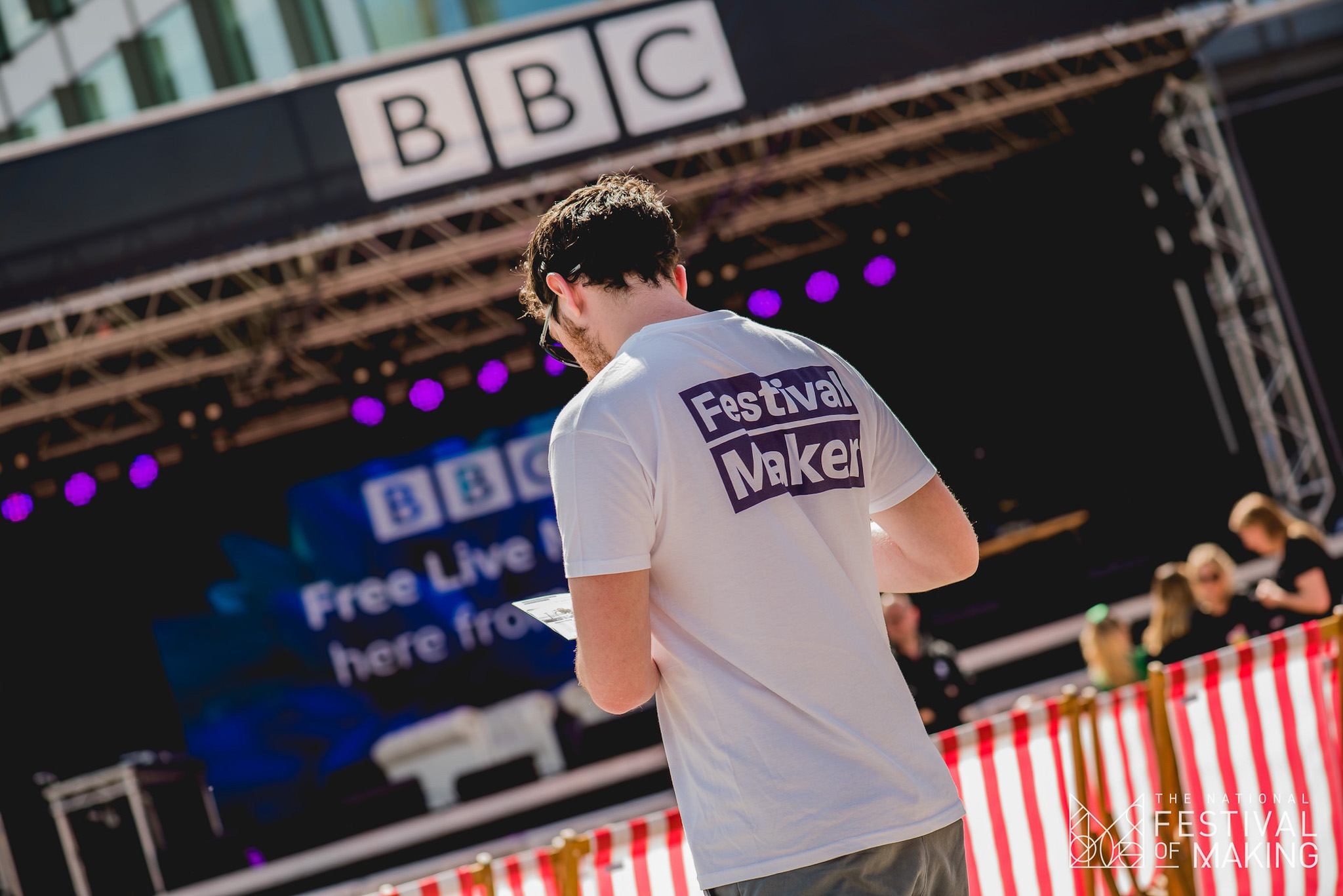 A person wearing a Festival Maker T-shirt stands in front of a stage with a BBC sign. The stage has bright lights, and there are red-and-white striped barriers in the foreground.