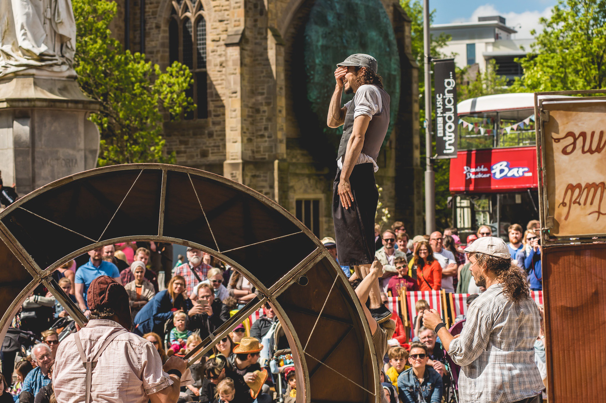 A street performer stands on a large wheel, covering his face, while a crowd watches outdoors. Two other performers are nearby, and a church is visible in the background.