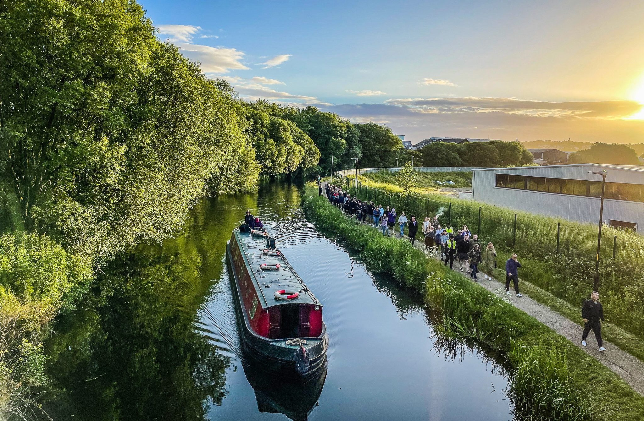 A narrowboat travels along a tree-lined canal at sunset, with a large group of people walking on the towpath beside it. The sky is clear with the sun low on the horizon.