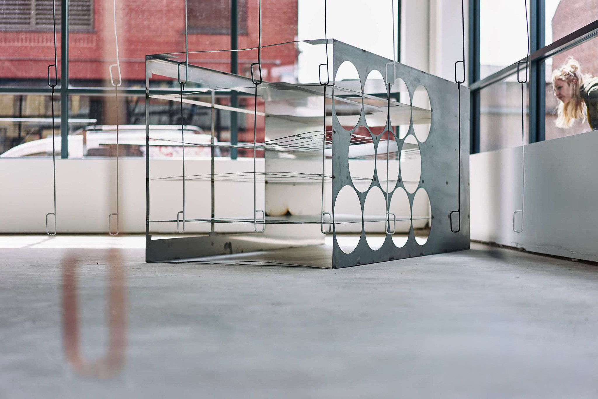 A metal sculpture circular cut-outs sits on a concrete floor in a sunlit room with large glass windows. A person with blonde hair is visible through the window in the background.