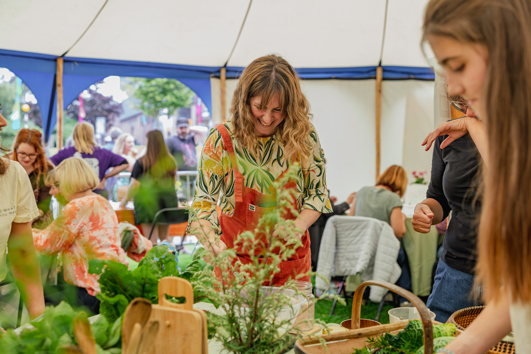 A group of people, mostly women, gather round a table with various herbs and plants inside a marquee, taking part in a practical activity. One woman in the centre smiles as she works with the greenery.