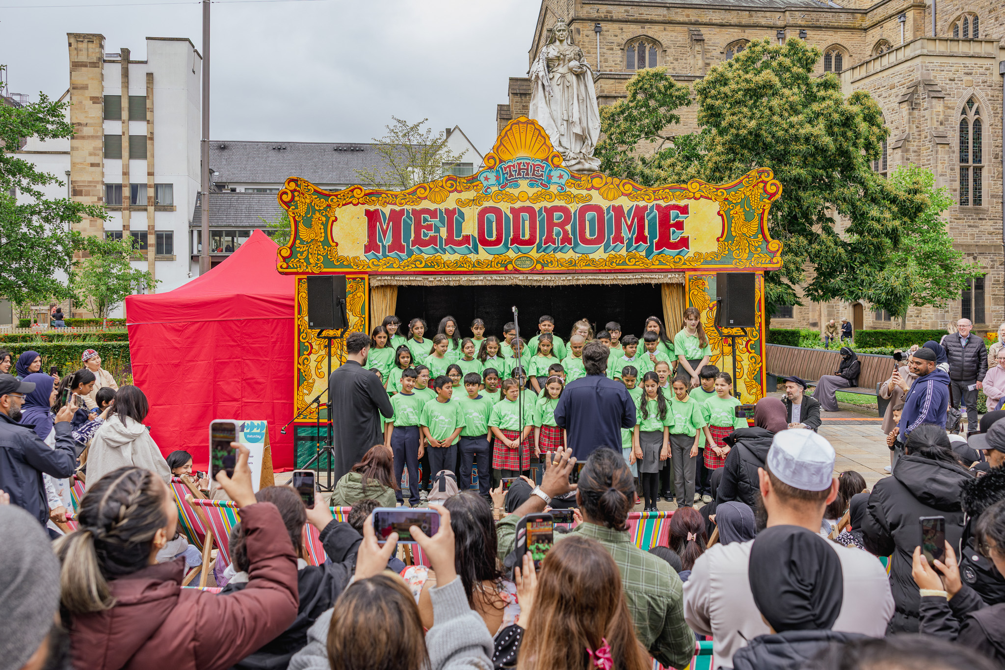 A group of children in green shirts sing on an outdoor stage labelled Melodrome as a crowd watches, many taking photos. The event takes place in a city square with historic buildings in the background.