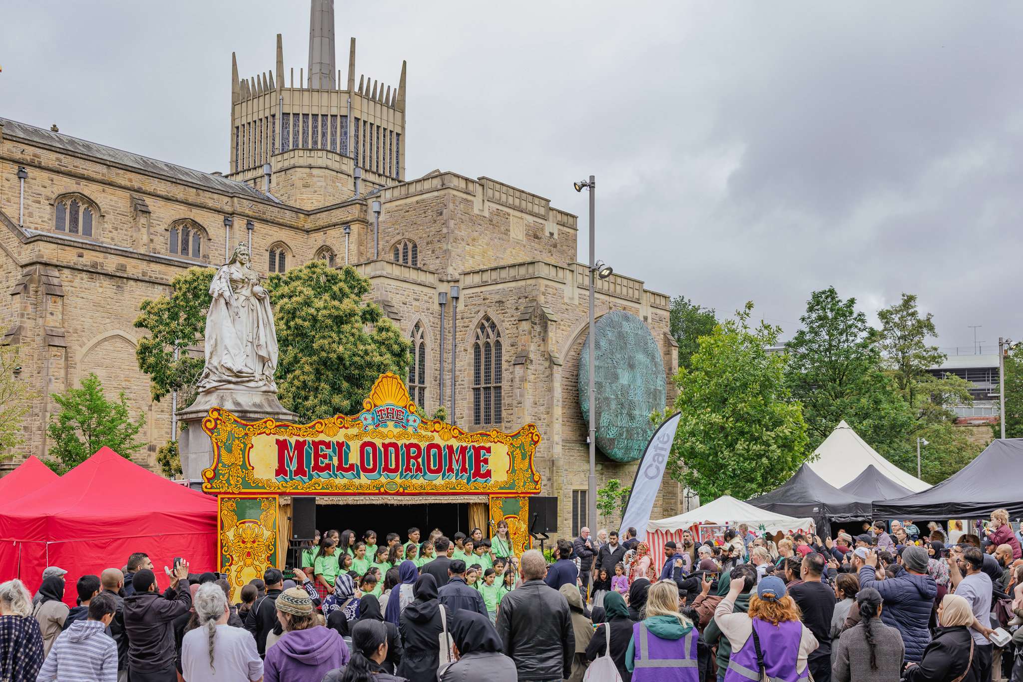 Children in green shirts sing on an outdoor stage labeled “Melodrome” as a crowd watches and takes photos, set in a city square with historic buildings in the background.