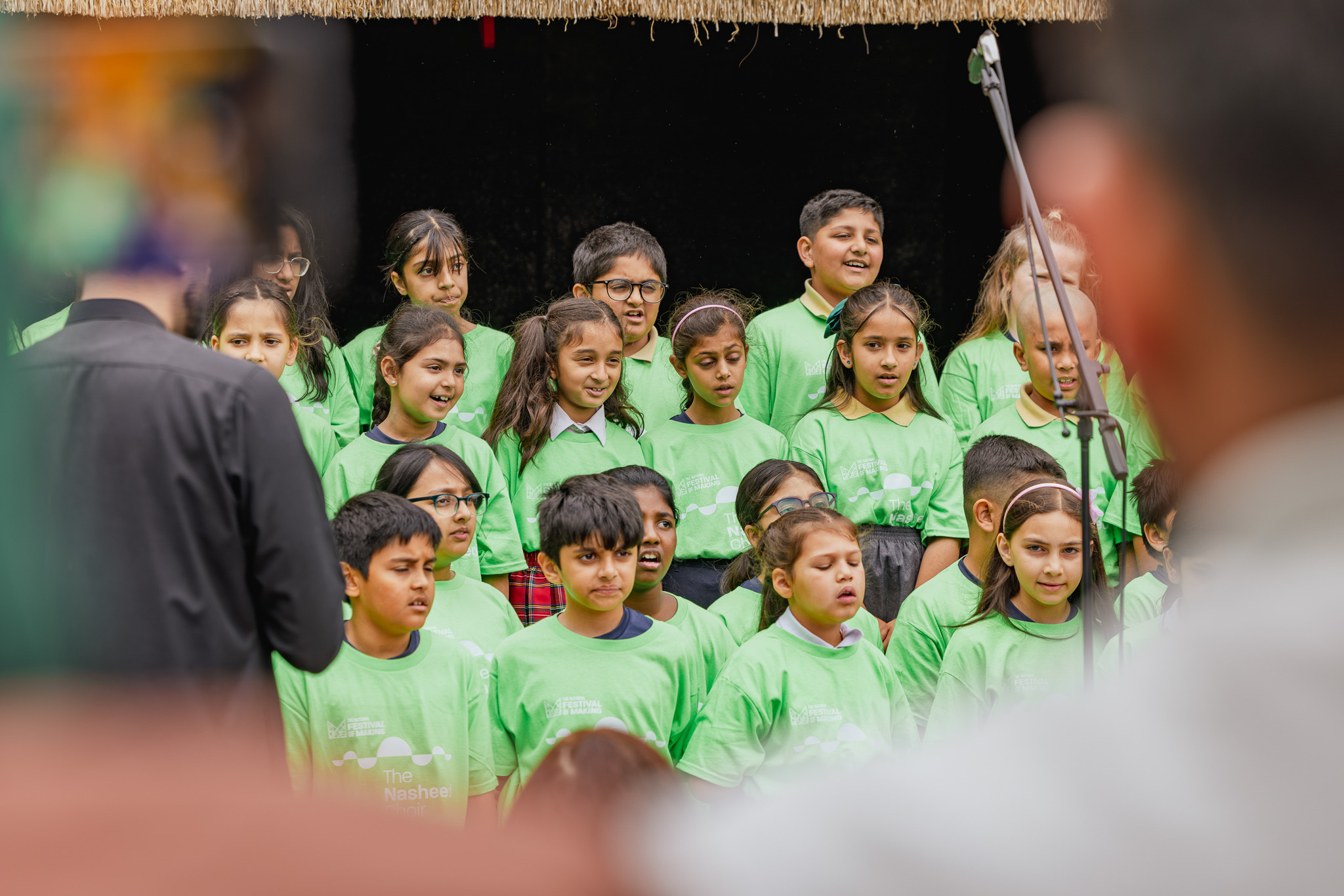 A group of children wearing matching green T-shirts stand together, singing or performing on stage. Some adults are partially visible in the foreground, and the background is dark.