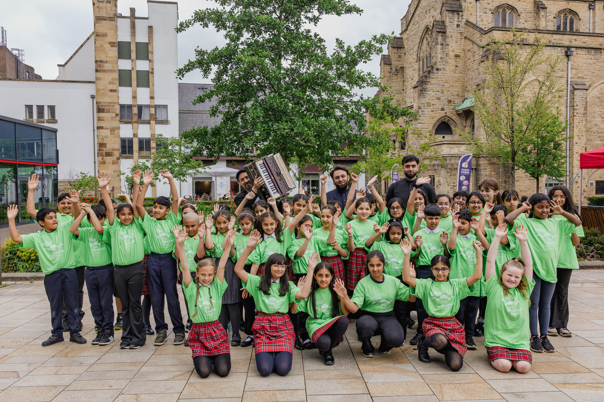 Cheerful children in green shirts and plaid skirts stand outside with raised arms, posing joyfully, while two men hold musical instruments in a lively plaza beside historic stone buildings.