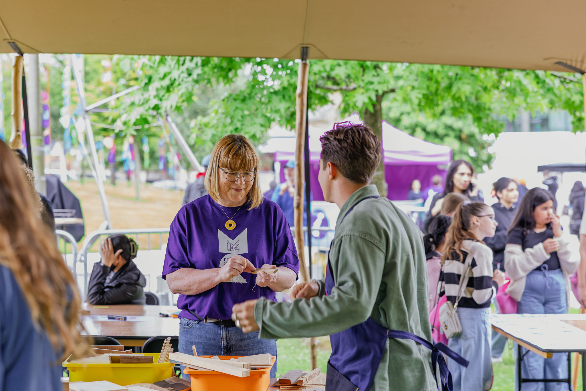 A volunteer in a purple shirt helps a visitor at an outdoor workshop, surrounded by people and children under a marquee with trees and activity stations in the background.