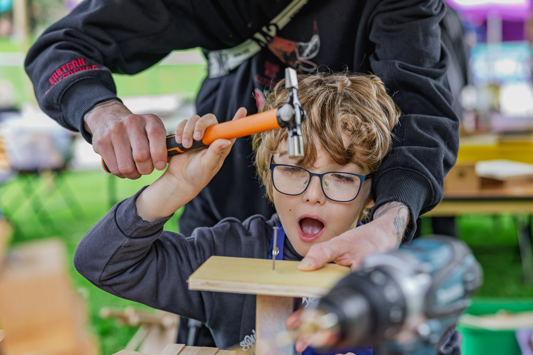 Child with glasses hammers a nail into wood, guided by an adult's hands, in a outdoor setting.
