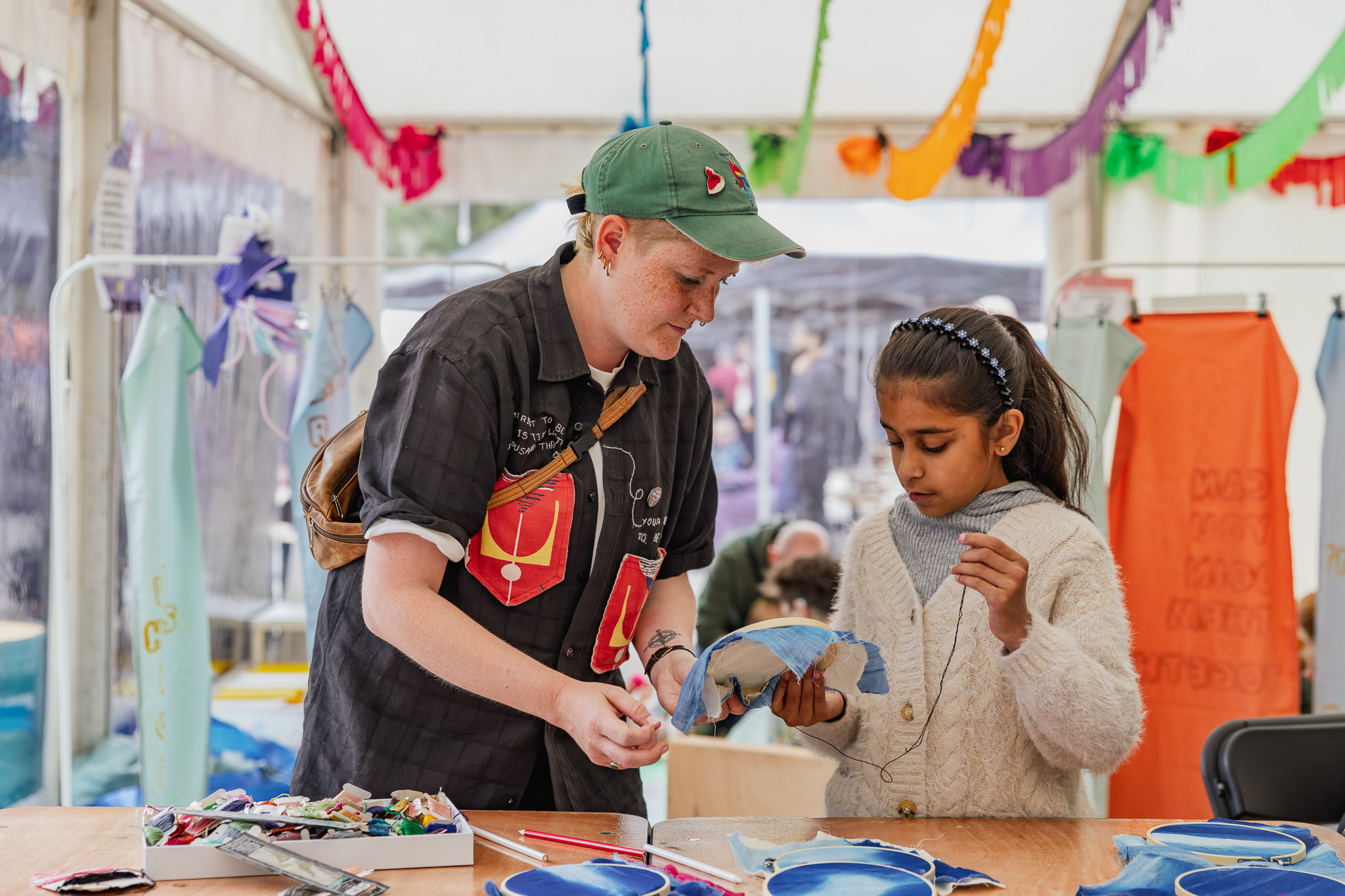 An adult helps a young girl with a craft project inside a colourful tent. The girl is sewing fabric, whilst the adult offers guidance. Craft supplies are spread on the table, and colourful decorations hang above.