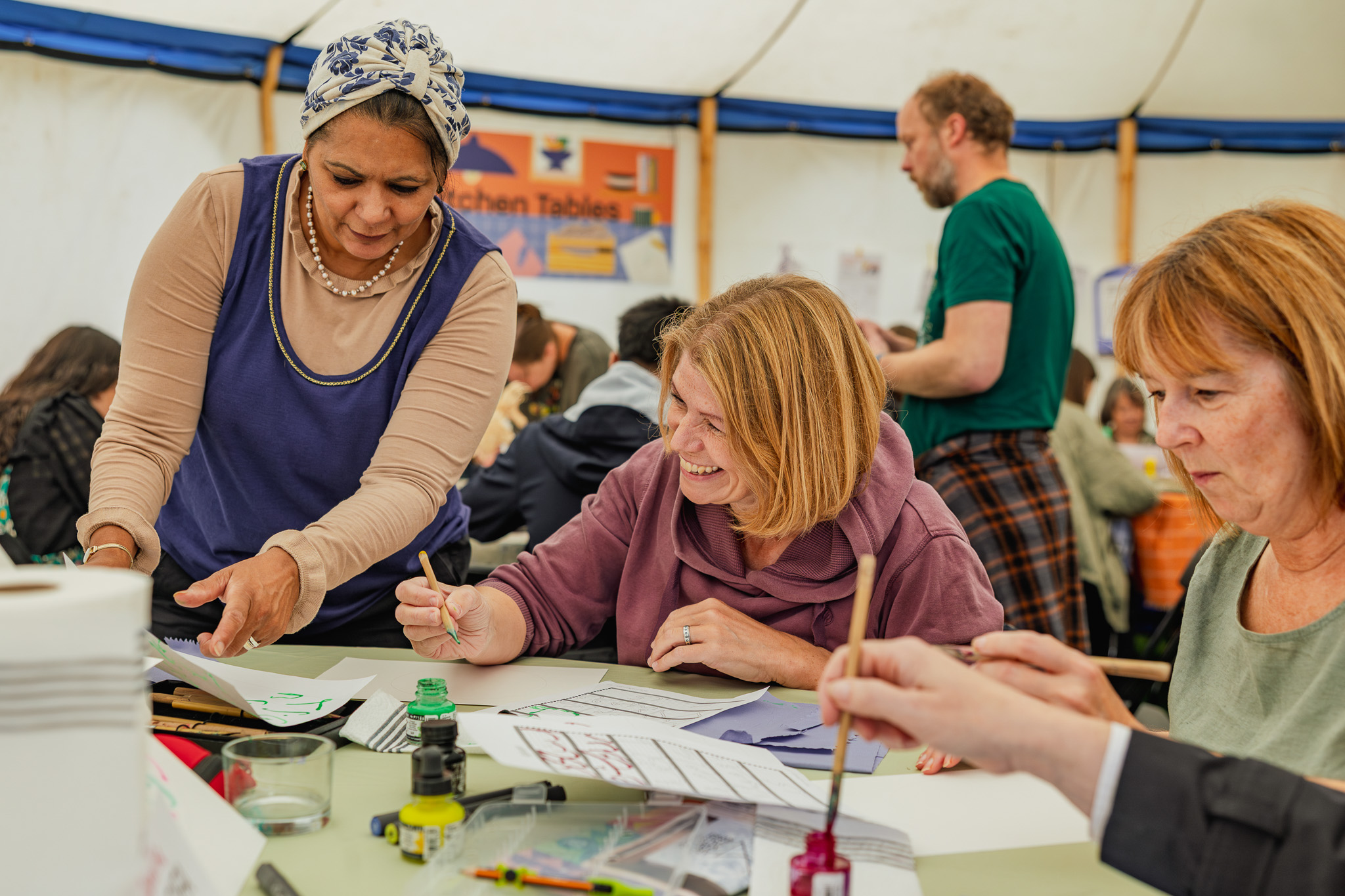 A group of adults work together at a table, drawing and writing with art supplies. One woman stands and assists others, who are seated and smiling inside a busy, well-lit marquee.