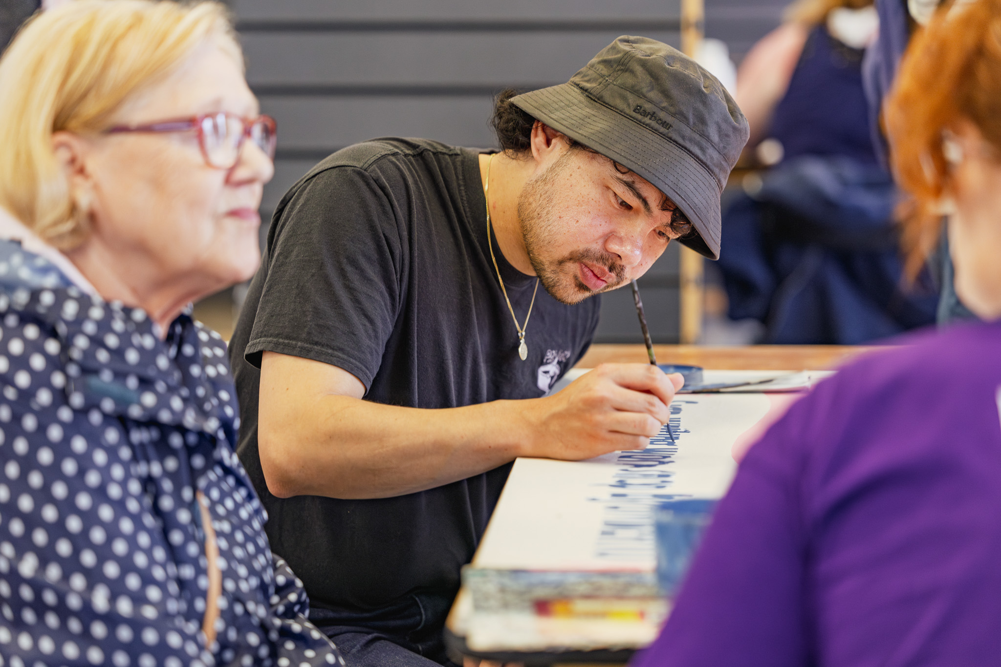 A man wearing a black hat and T-shirt is focused on painting blue lines on a white canvas at a table, whilst two women sit nearby, one in a polka dot jacket and the other in purple.