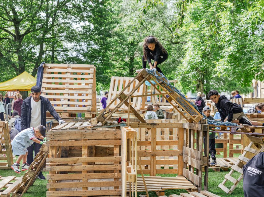Children and adults build and climb on wooden pallet structures in a park surrounded by green trees. People work together creatively, using ropes and planks, at an outdoor event with a lively, playful atmosphere.