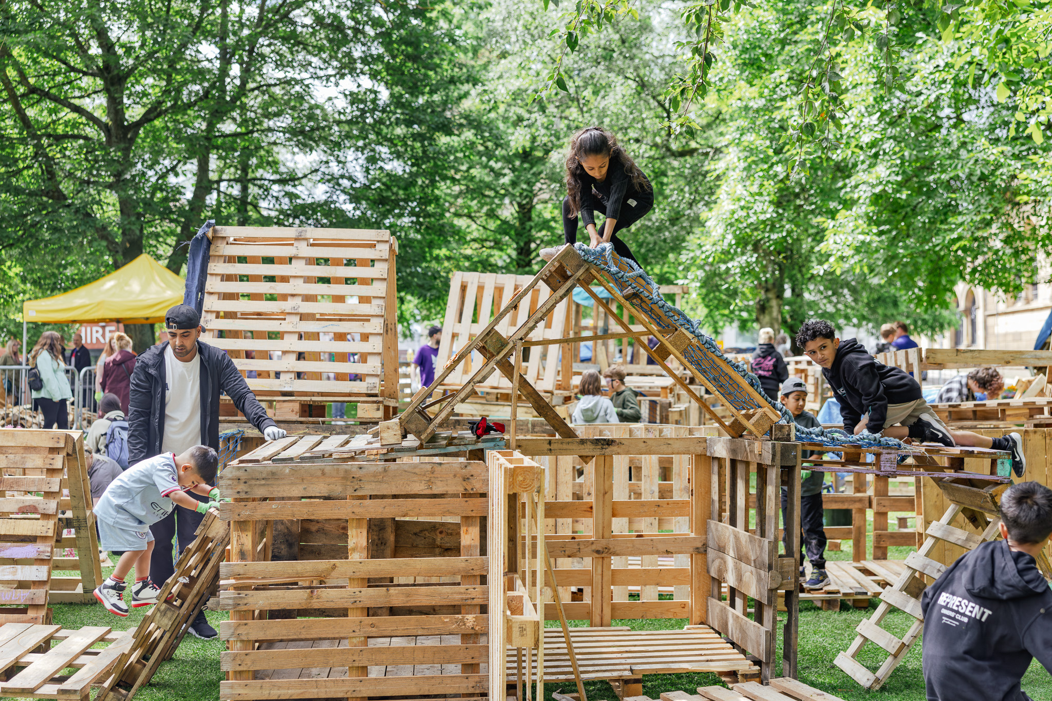 Children and adults build and climb on wooden pallet structures in a park surrounded by green trees. People work together creatively, using ropes and planks, at an outdoor event with a lively, playful atmosphere.
