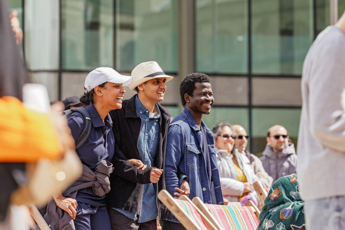 A group of people outdoors, smiling and standing together. Three people are in the foreground, dressed casually in hats and jackets, while others watch in the background in front of a modern glass building.