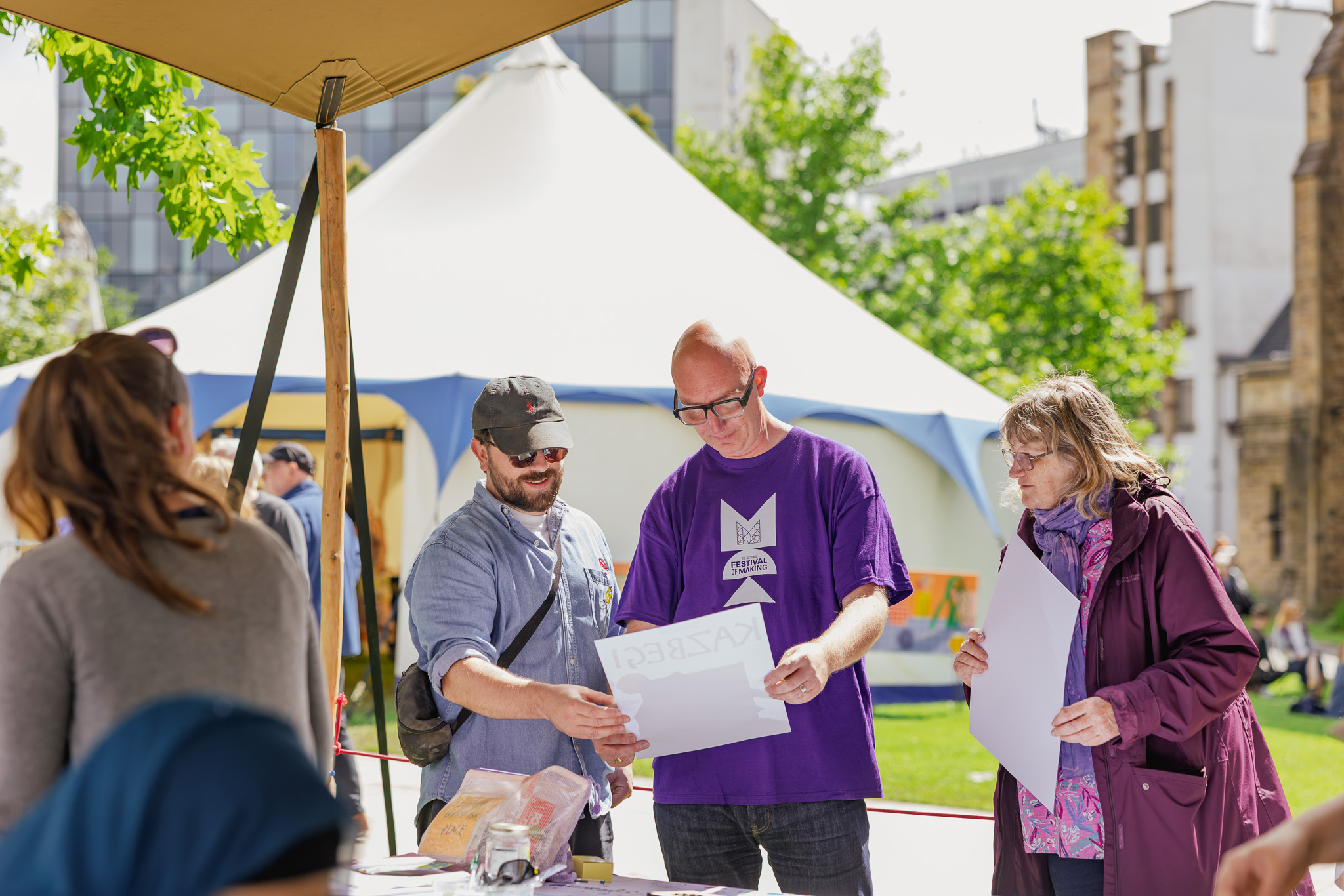 Three people stand outdoors at a stall, one holding a poster and wearing a purple shirt, whilst others look on and discuss. A large white marquee and trees are visible in the background on a sunny day.
