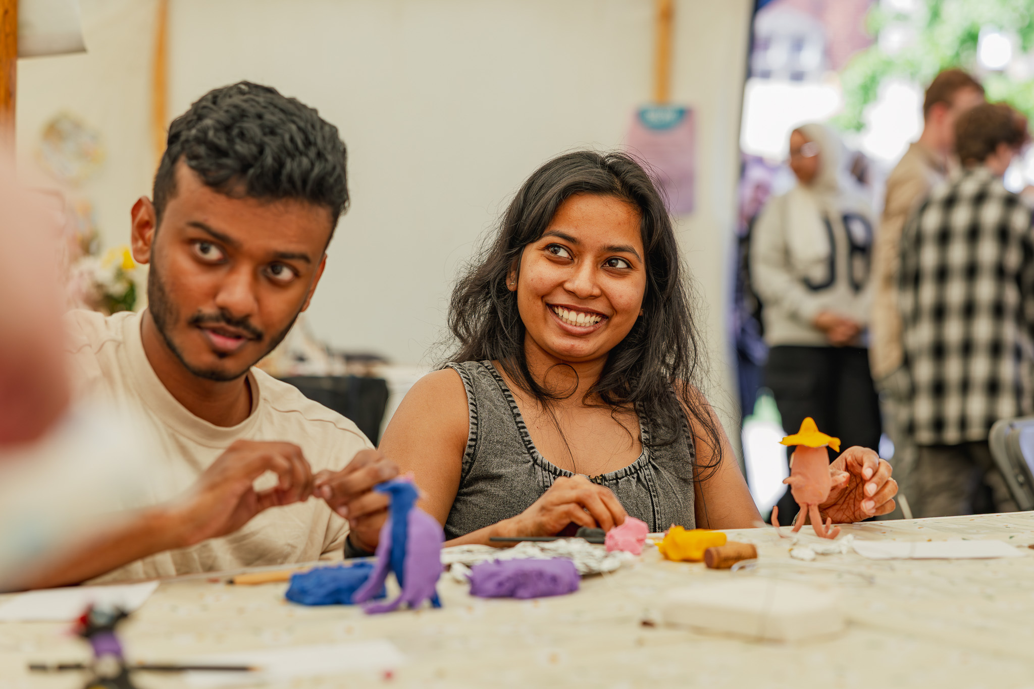 Two people smile and sculpt colourful clay figures at a table indoors, with more people and craft materials visible in the background.