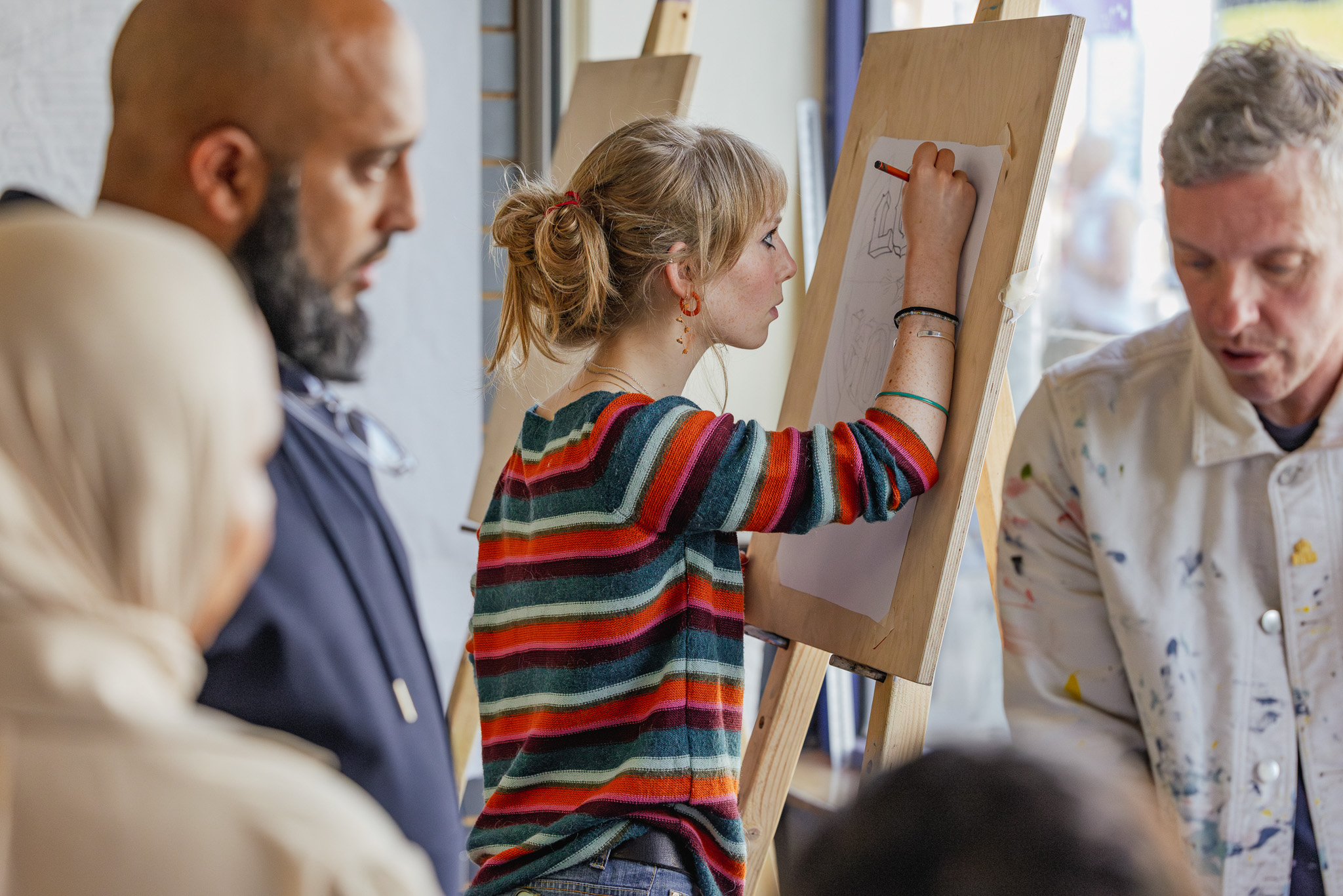 A woman in a colourful striped jumper draws on a large sheet of paper at an easel, surrounded by other adults who are watching and participating in an indoor art class.