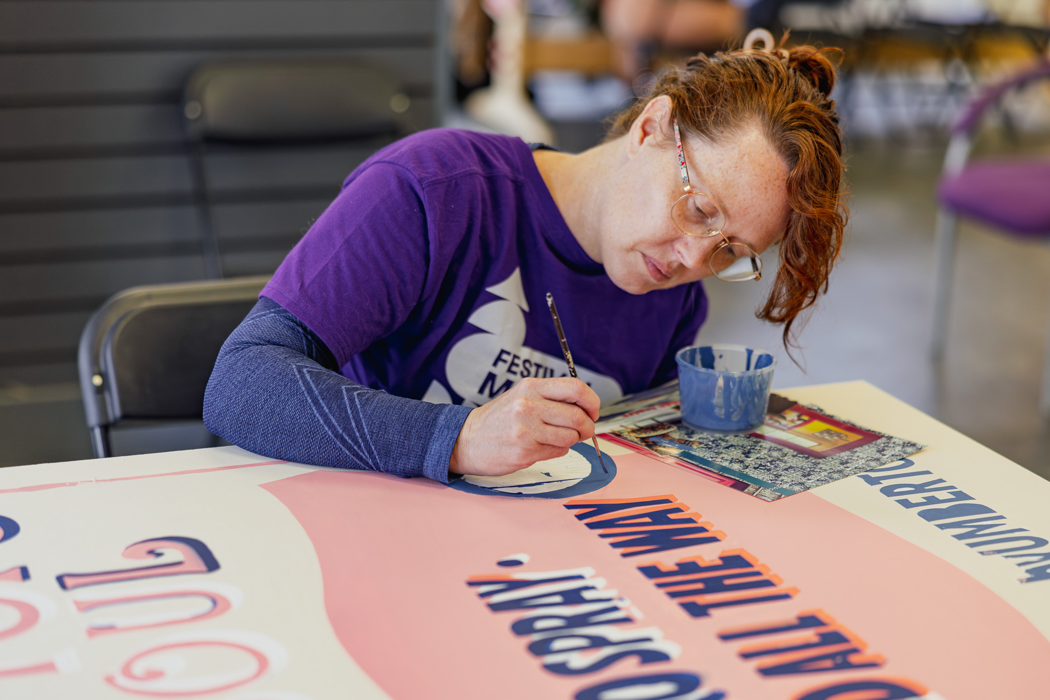 A person with ginger hair and glasses, wearing a purple shirt, sits at a table painting letters on a large sign. They focus intently, holding a paintbrush near a cup of blue paint, with art supplies nearby.