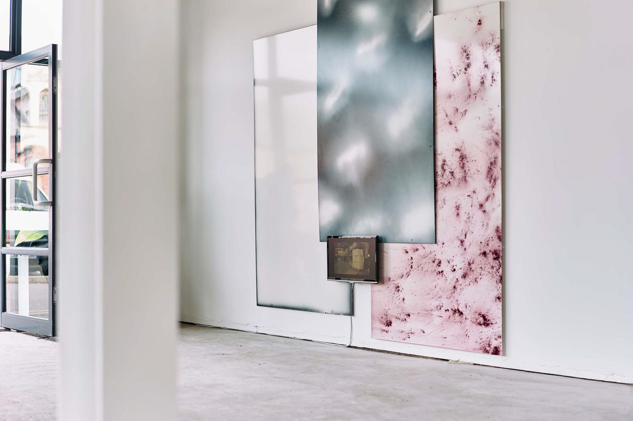 Three large abstract panels—one white, one metallic grey, and one pink with marbled texture—are propped against a white wall in a minimalist, empty room with concrete floors and a glass door.