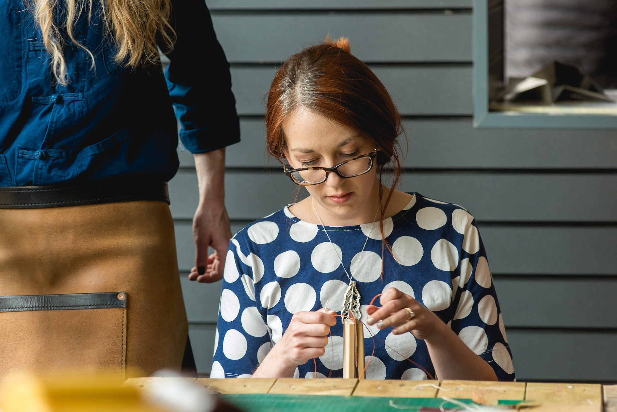 A woman with glasses, wearing a navy blue shirt with white polka dots, focuses on crafting with string at a table. A person in a blue shirt and brown apron stands nearby. They are indoors by a grey panelled wall.