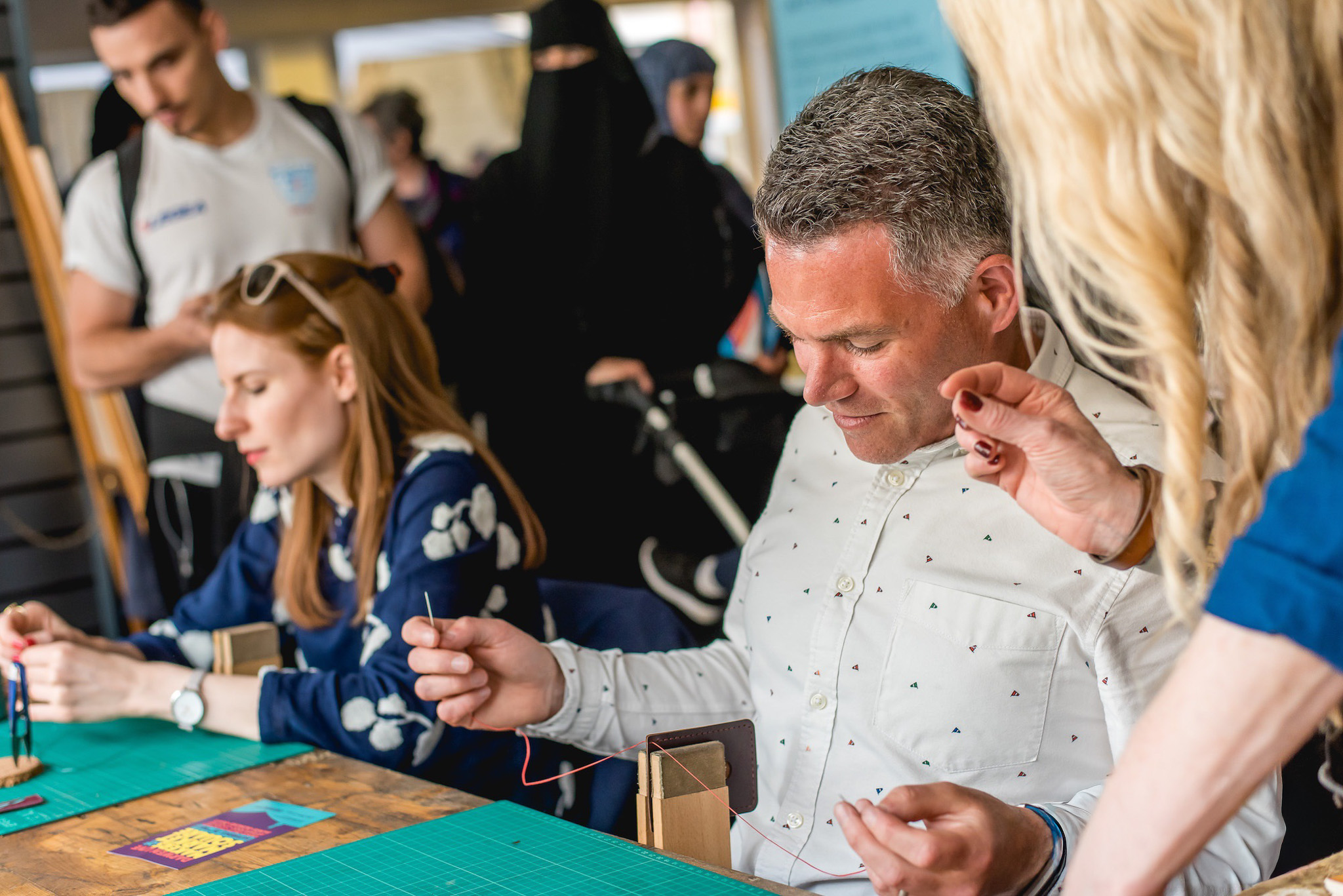 A group of people engaged in a workshop. A man in a white shirt and a woman are sewing.
