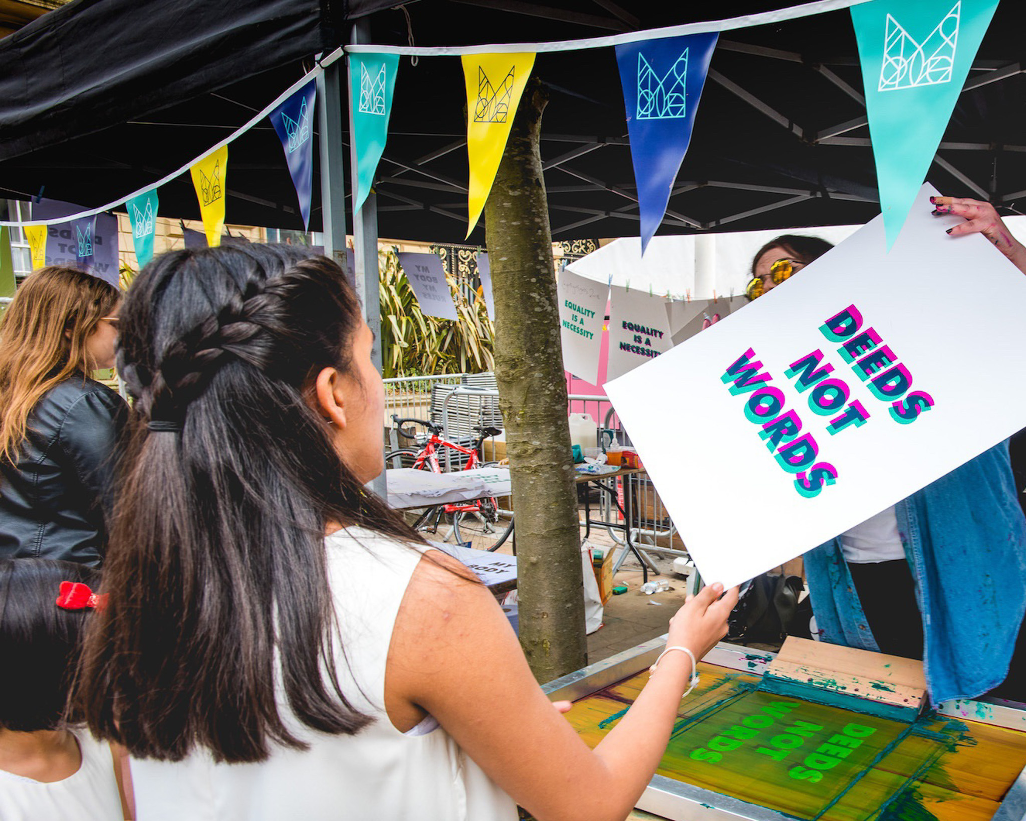 A person holds a sign reading DEEDS NOT WORDS at an outdoor event stall decorated with colourful bunting. Another person stands nearby, watching. Tables and art materials are visible in the background.