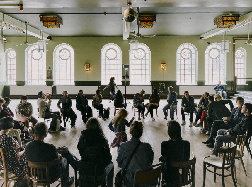 A group sits in a large circle of chairs in a spacious room with arched windows and wooden floors, watching a performance as a child dances in the centre.
