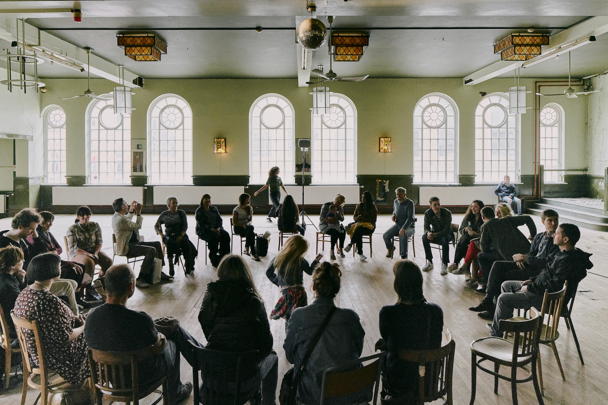 A group sits in a large circle of chairs in a spacious room with arched windows and wooden floors, watching a performance as a child dances in the centre.