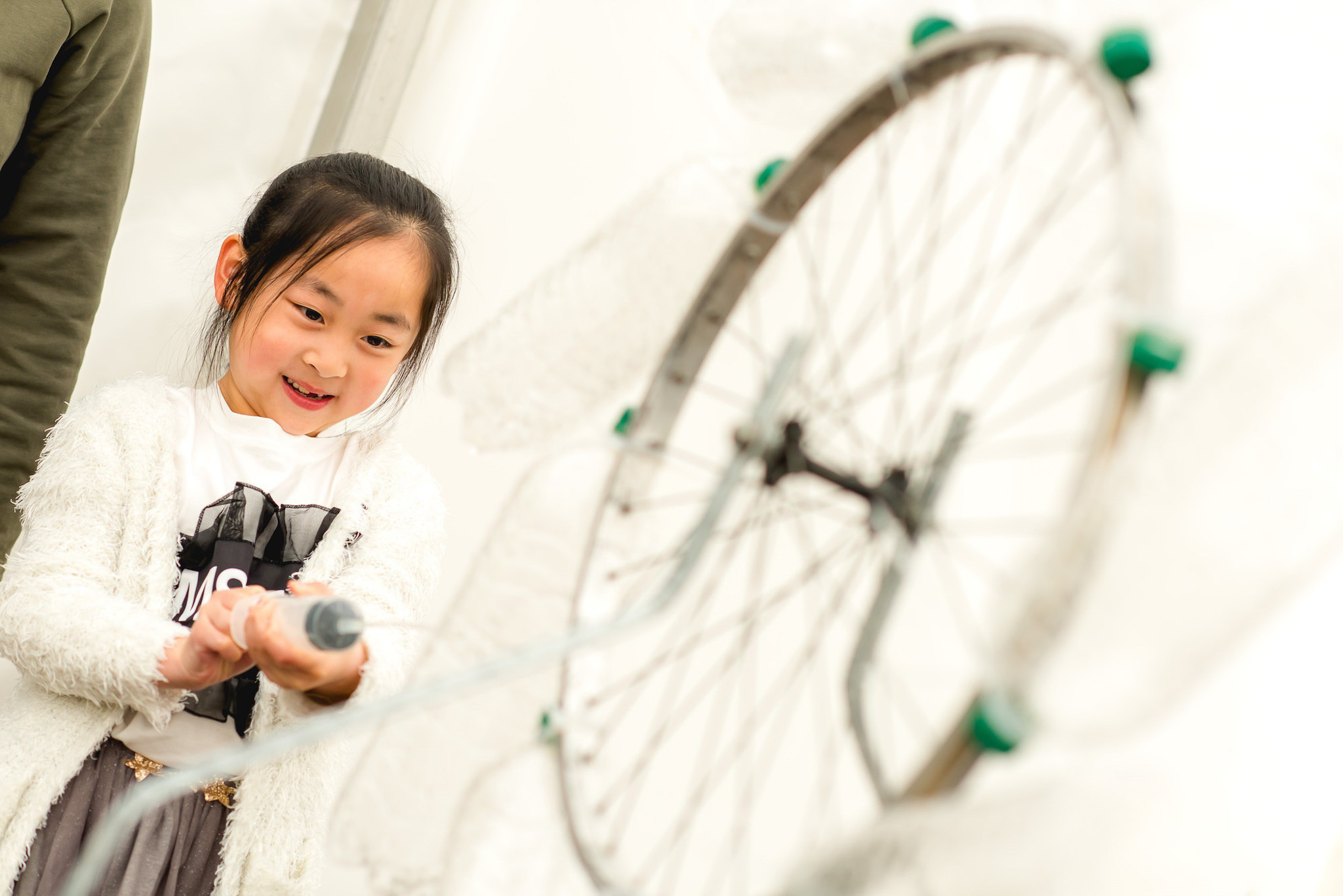 A young girl smiles as she interacts with a spinning bicycle wheel contraption, using a tool or handle attached to it. The background is bright and minimal.