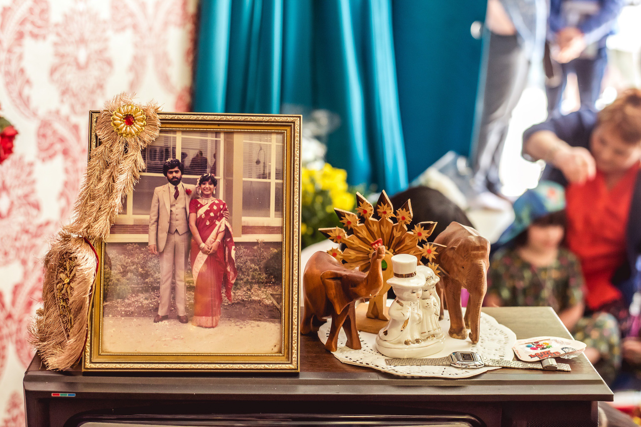 A framed vintage photo of a couple stands on a table with wooden elephant figurines, a ceramic couple, a small bouquet, and other decorative items; people and bright teal curtains are blurred in the background.
