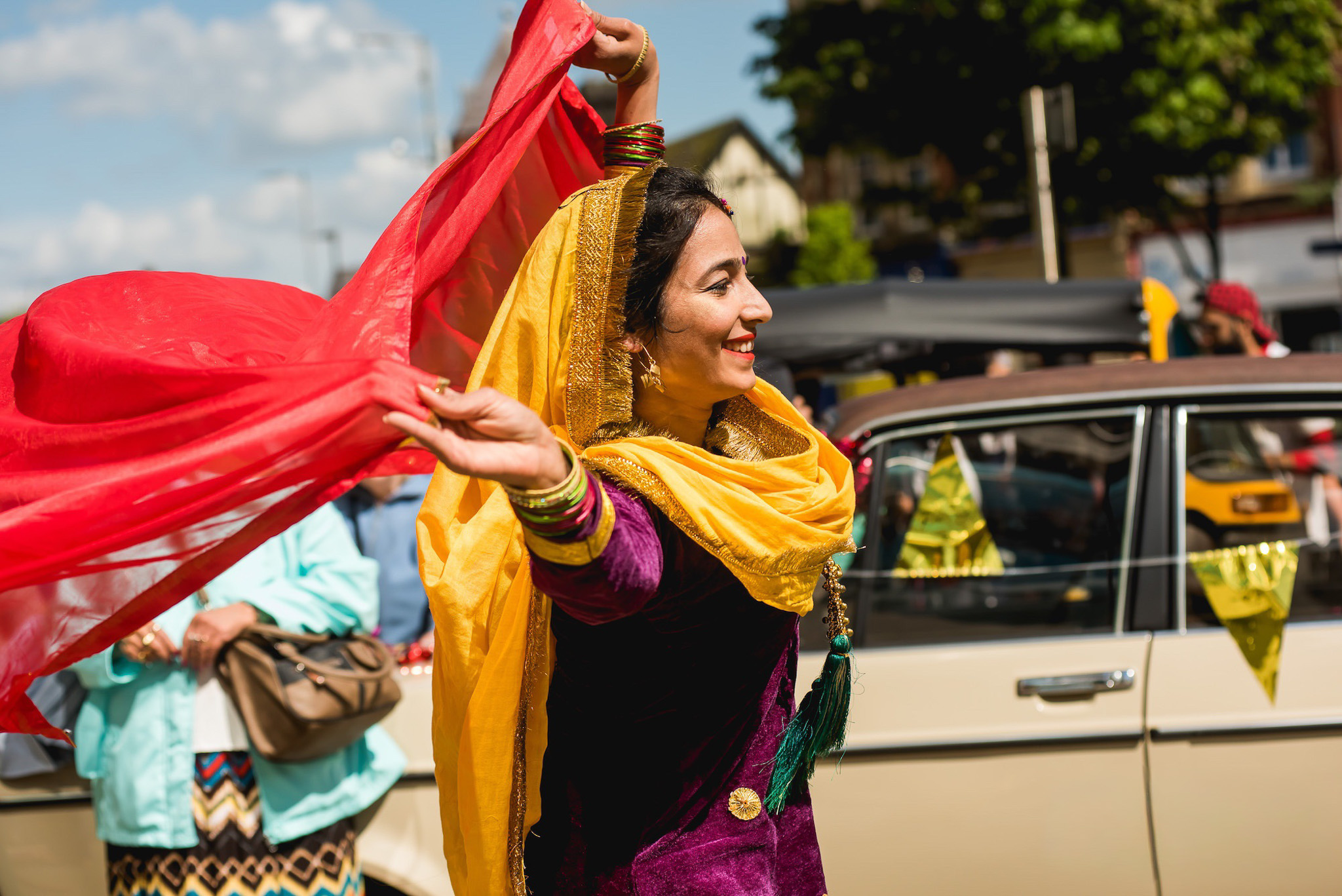 A woman wearing traditional clothing and jewellery dances joyfully outdoors, holding a red scarf above her head. She is smiling, with a yellow scarf draped over her shoulders, and people and a vintage car are visible in the background.
