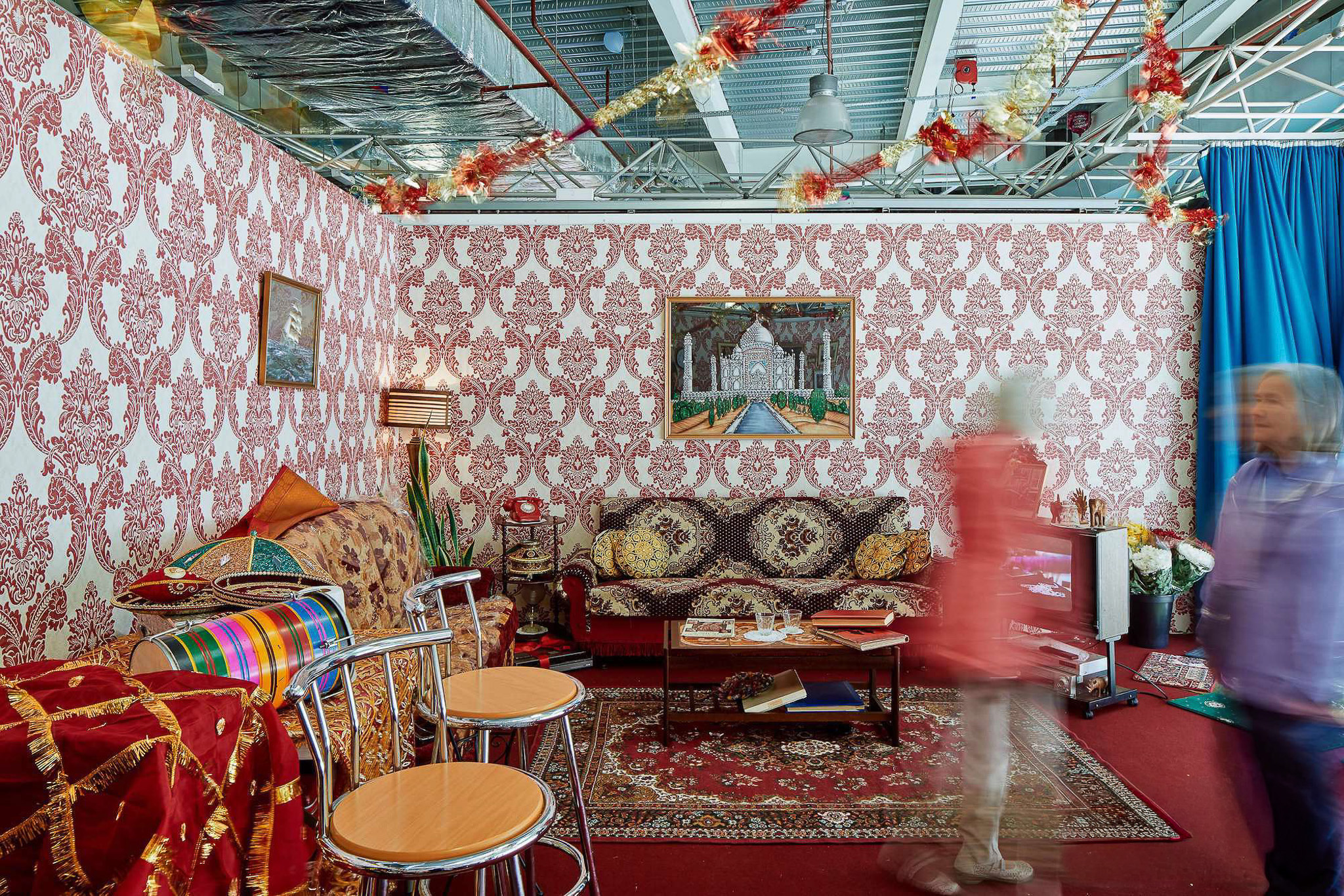 A room with ornate red and white wallpaper, antique furniture, patterned rugs, framed art, and festive decorations. Two people walk by, blurred in motion. The ceiling shows exposed pipes and lights.