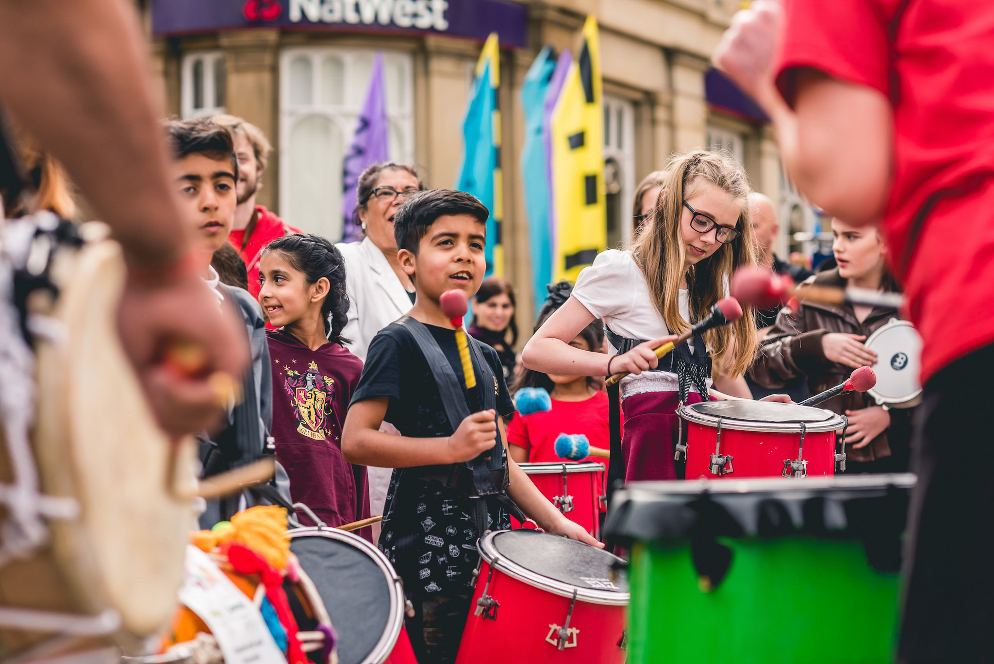 Children and adults play colourful drums at an outdoor street event, with vibrant flags and a NatWest bank in the background. The group looks focused and joyful as they perform together.