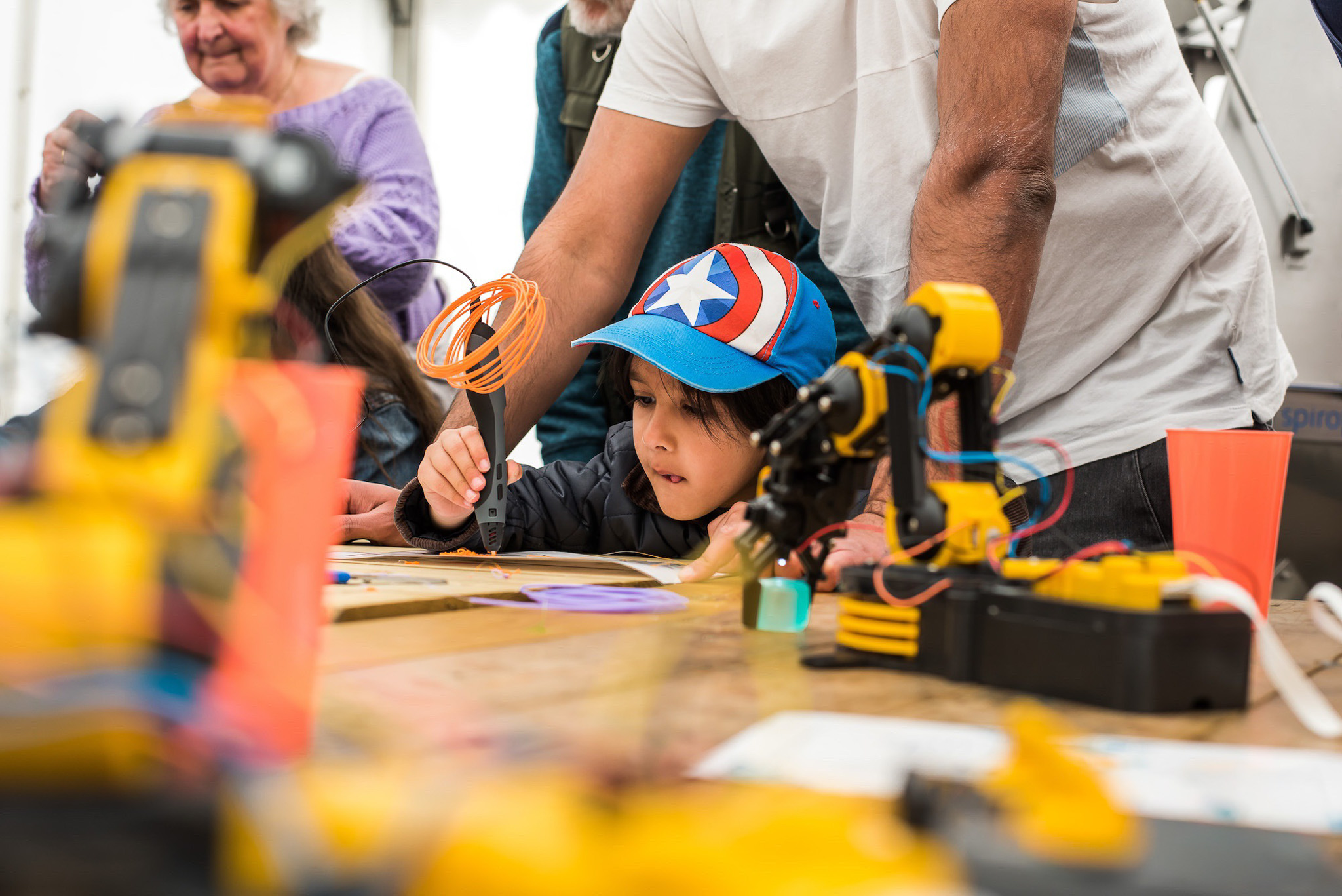 A young child in a superhero cap uses a 3D printing pen under adult guidance. Nearby, robotic arms create a lively, experimental atmosphere.