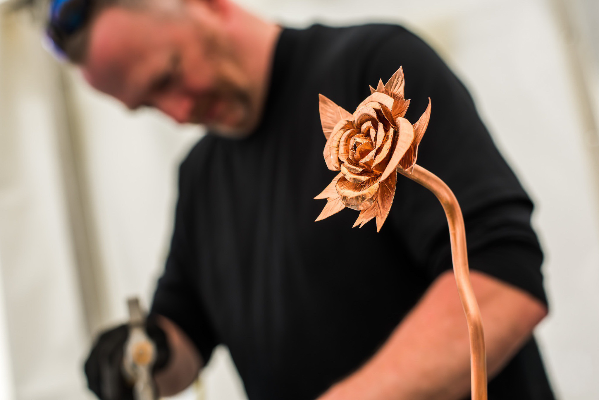 A close-up of a detailed copper rose sculpture in focus, with a blurred person in black working in the background.