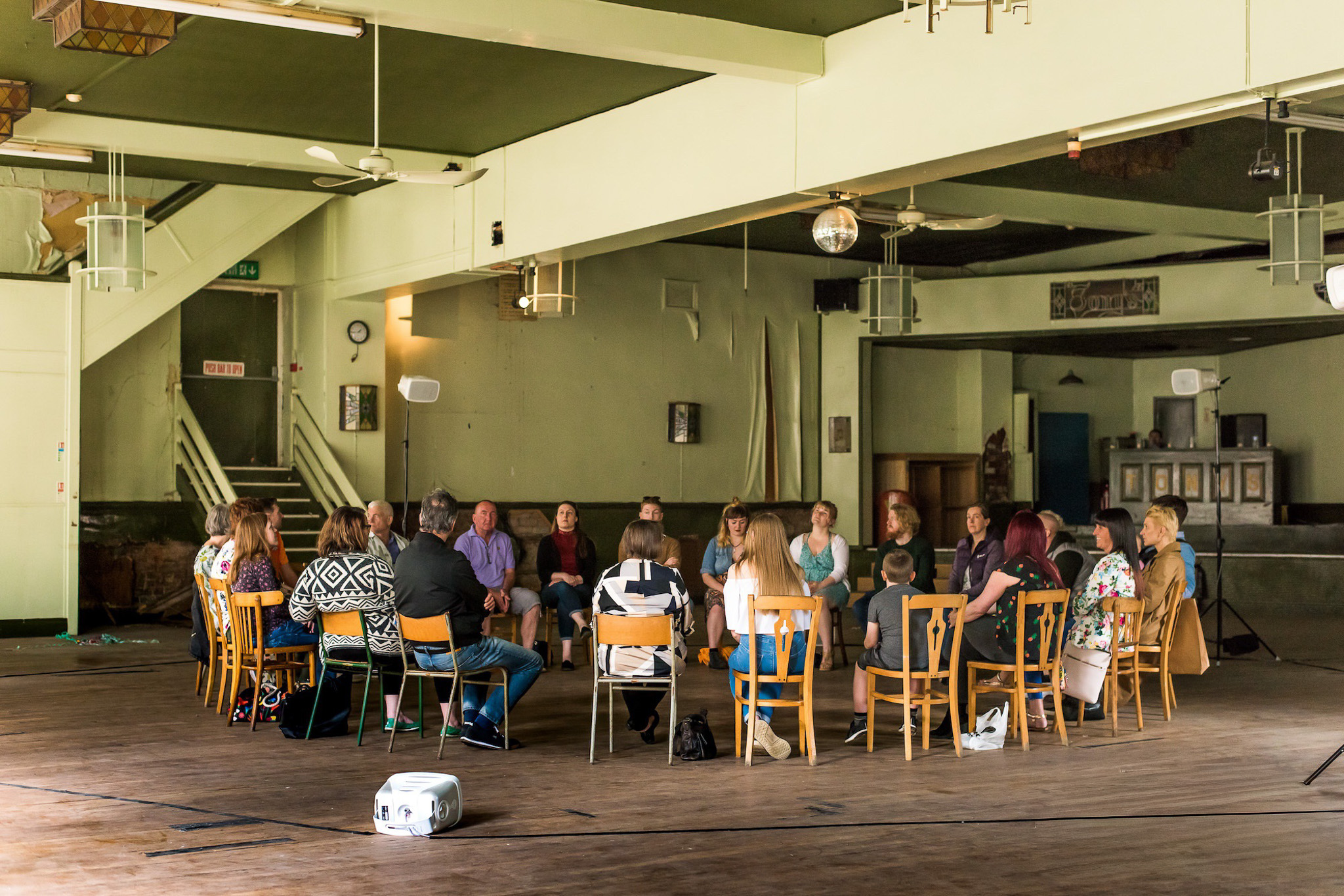 A group of people sit in a circle on wooden chairs in a large, spacious room with high ceilings, wooden floors, and minimal decoration, suggesting a meeting, workshop, or group discussion in progress.