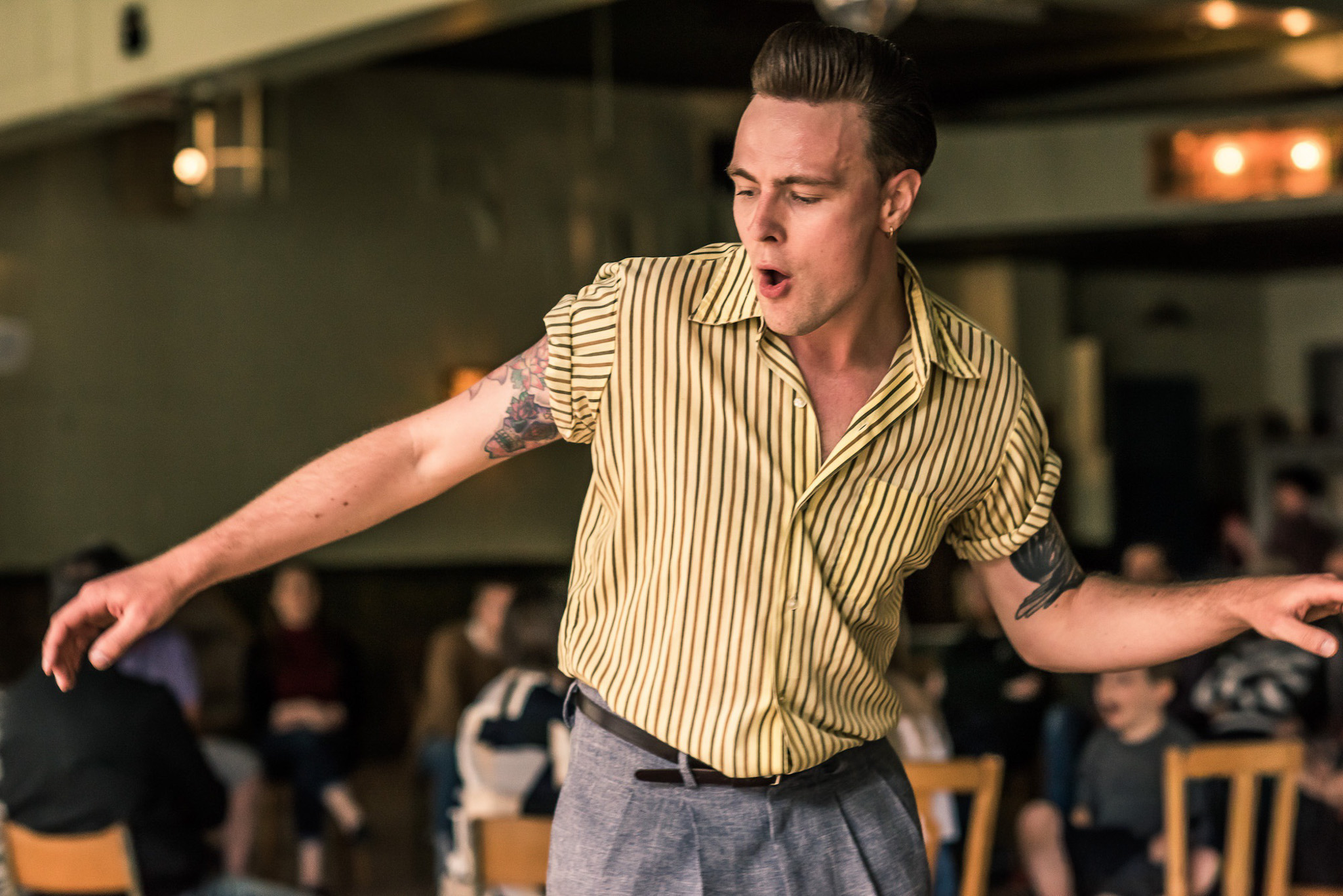 A man in a yellow striped shirt and grey trousers dances energetically indoors, with his arms extended. People are seated in the background, observing. The setting appears lively and casual.