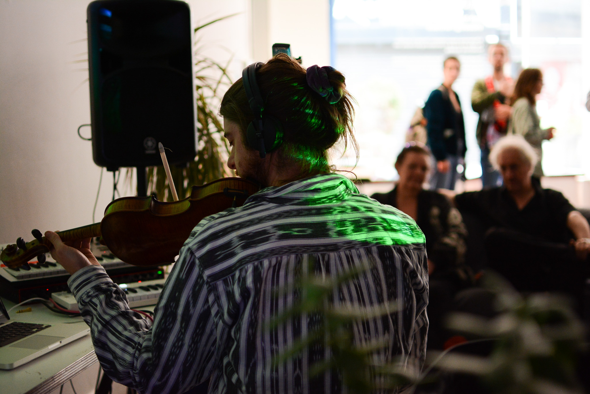 A person with headphones plays a violin in a room with electronic equipment, facing a small seated audience. Green light highlights their hair, and a few people stand in the background near a window.
