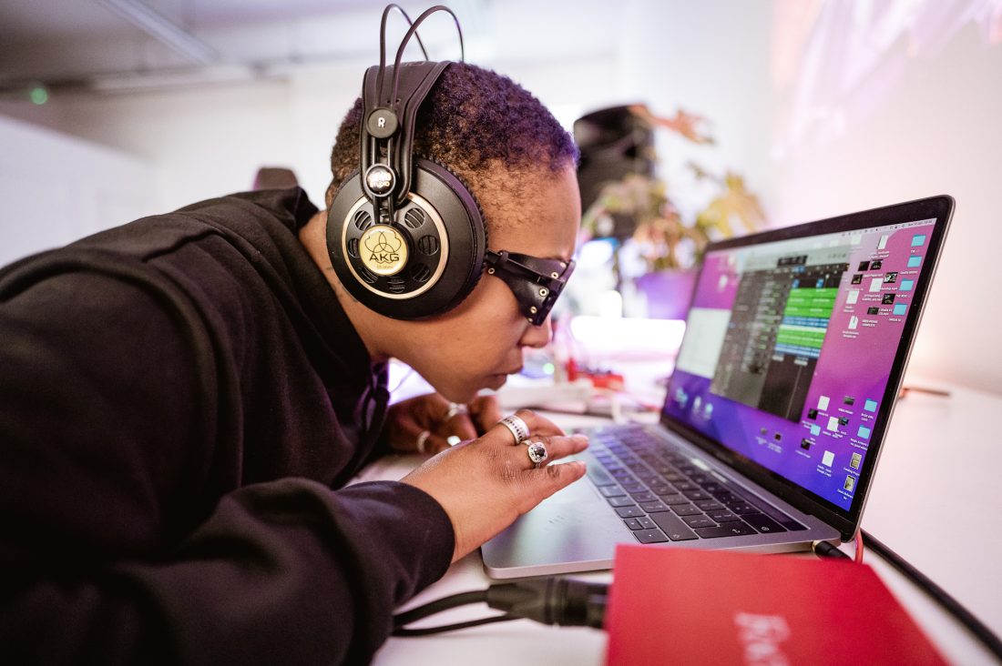A person wearing headphones and sunglasses leans closely towards a laptop screen, focusing intently whilst using the keyboard. The laptop displays audio editing software, and there is studio equipment nearby.