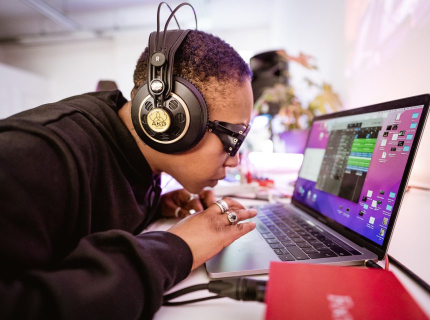 A person wearing headphones and sunglasses leans closely towards a laptop screen, focusing intently whilst using the keyboard. The laptop displays audio editing software, and there is studio equipment nearby.